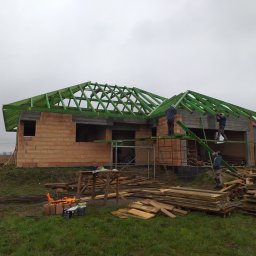 FENIX Paweł Oczkowski - Budowa domu jednorodzinnego w trakcie montażu zielonej więźby dachowej, widoczni pracownicy na rusztowaniach, ceglany budynek w stanie surowym otwartym, ognisko palone na ziemi obok składowanego...