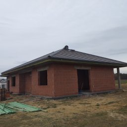 FENIX Paweł Oczkowski - Dom jednorodzinny w trakcie budowy: murowane ściany z czerwonej cegły, zamontowany ciemny dach z rynnami, widoczne palety i materiały budowlane na zewnątrz, pochmurne niebo.