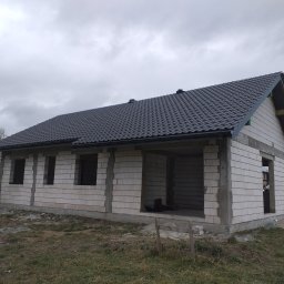 FENIX Paweł Oczkowski - Surowy stan domu jednorodzinnego z ciemną dachówką ceramiczną, widoczne niezadaszone otwory okienne i drzwiowe, szare niebo w tle.