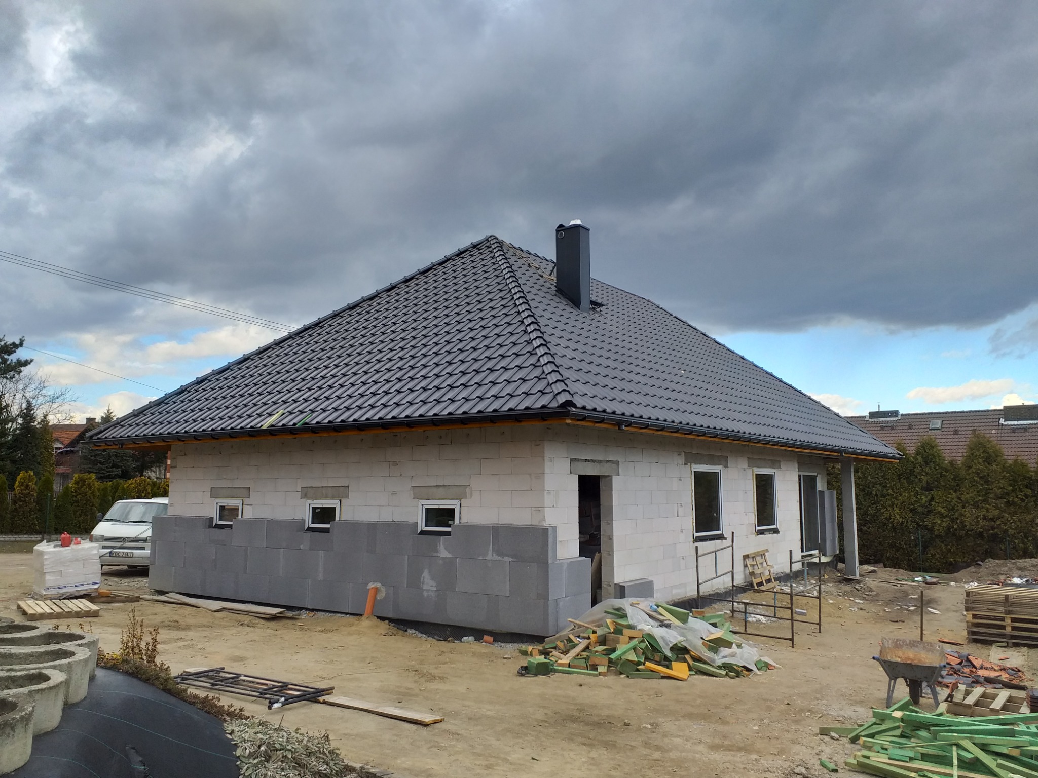 Dom jednorodzinny w trakcie budowy, widoczny z zewnątrz, z szarą dachówką i kominem, częściowo ocieplony, z widocznymi materiałami budowlanymi i narzędziami na placu.