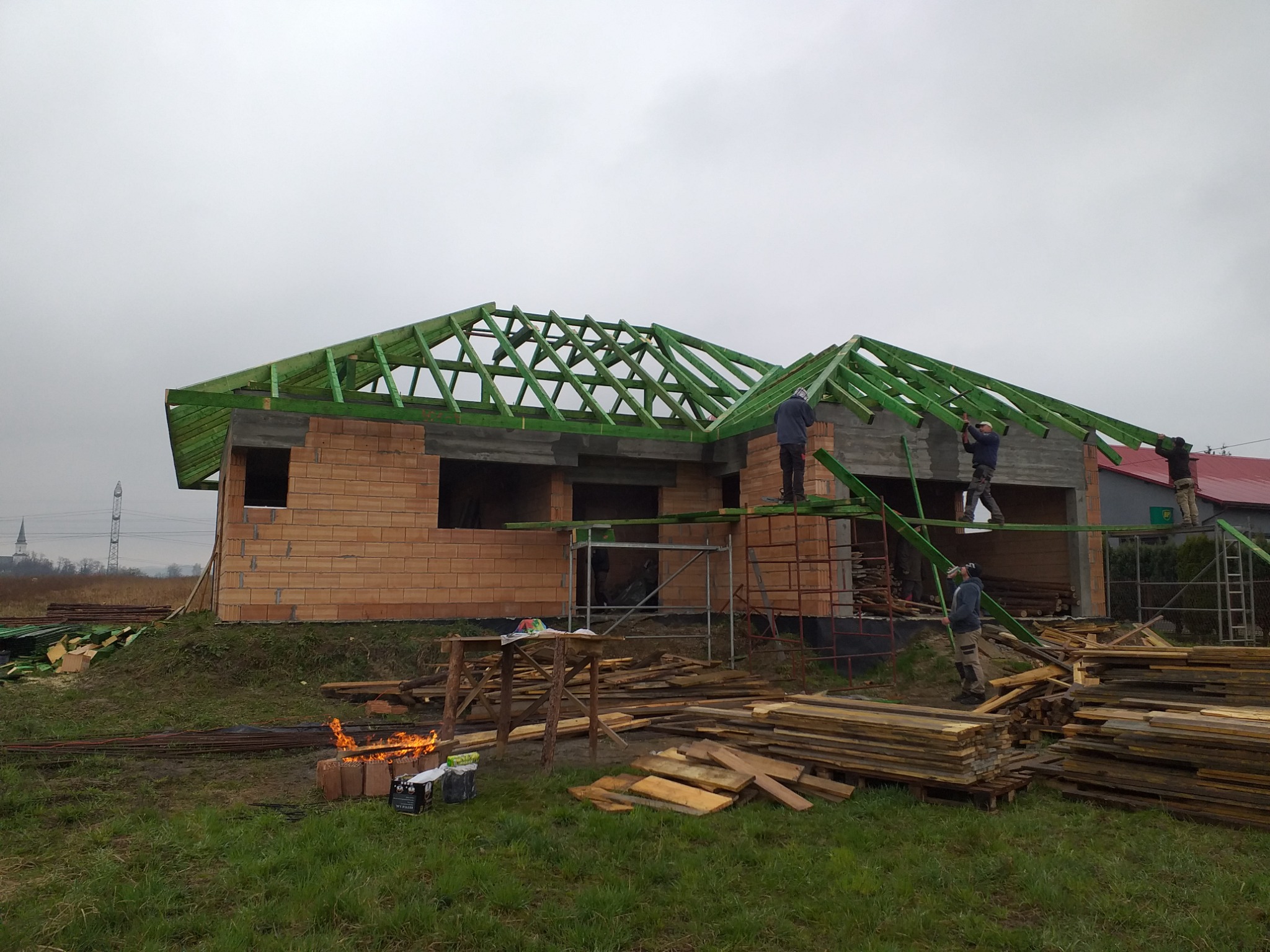 Budowa domu jednorodzinnego w trakcie montażu zielonej więźby dachowej, widoczni pracownicy na rusztowaniach, ceglany budynek w stanie surowym otwartym, ognisko palone na ziemi obok składowanego...