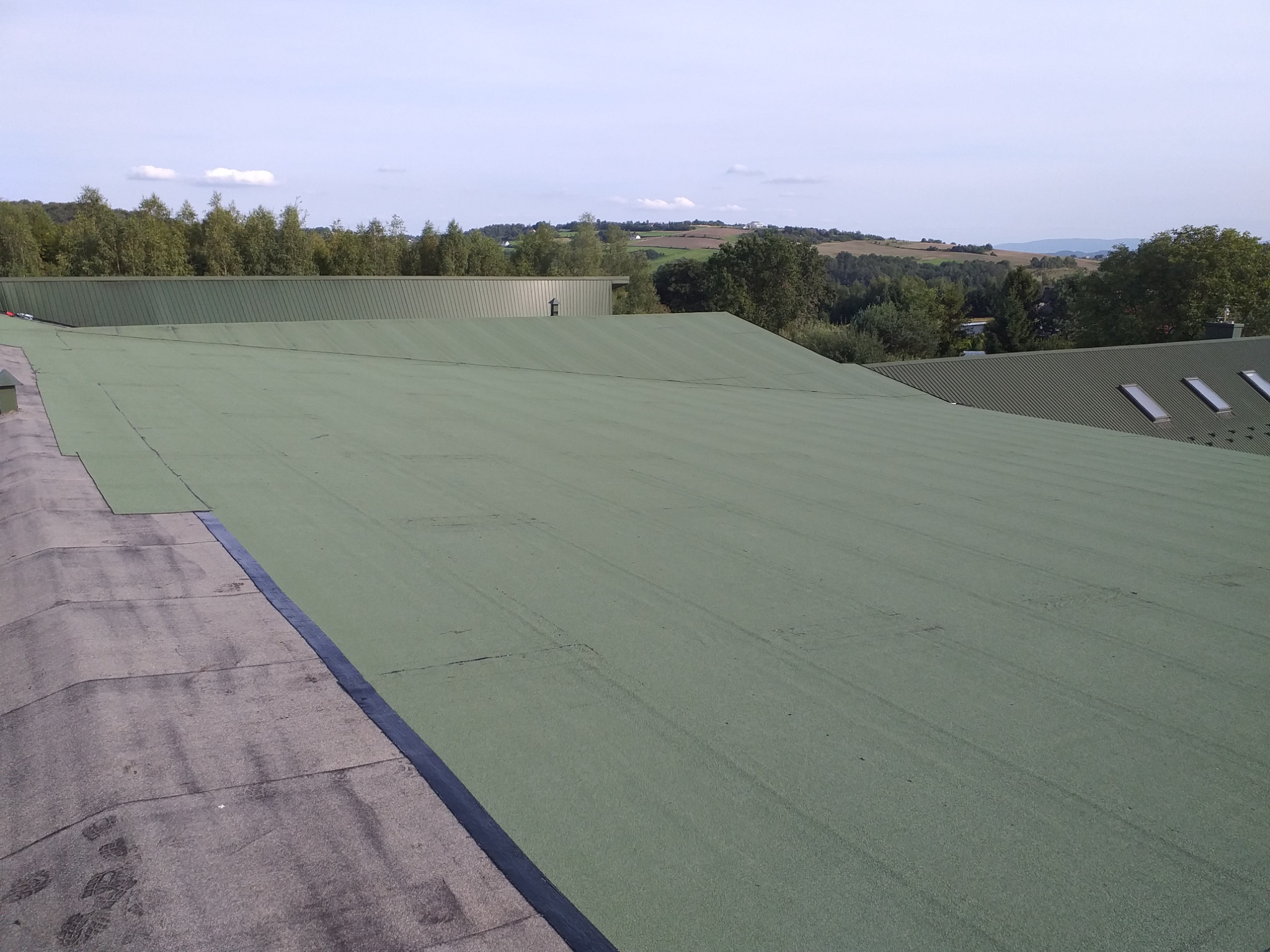 Rozległy, płaski dach pokryty zieloną papą, z widocznymi śladami stóp i fragmentami innych pokryć, na tle zielonych wzgórz i błękitnego nieba.
