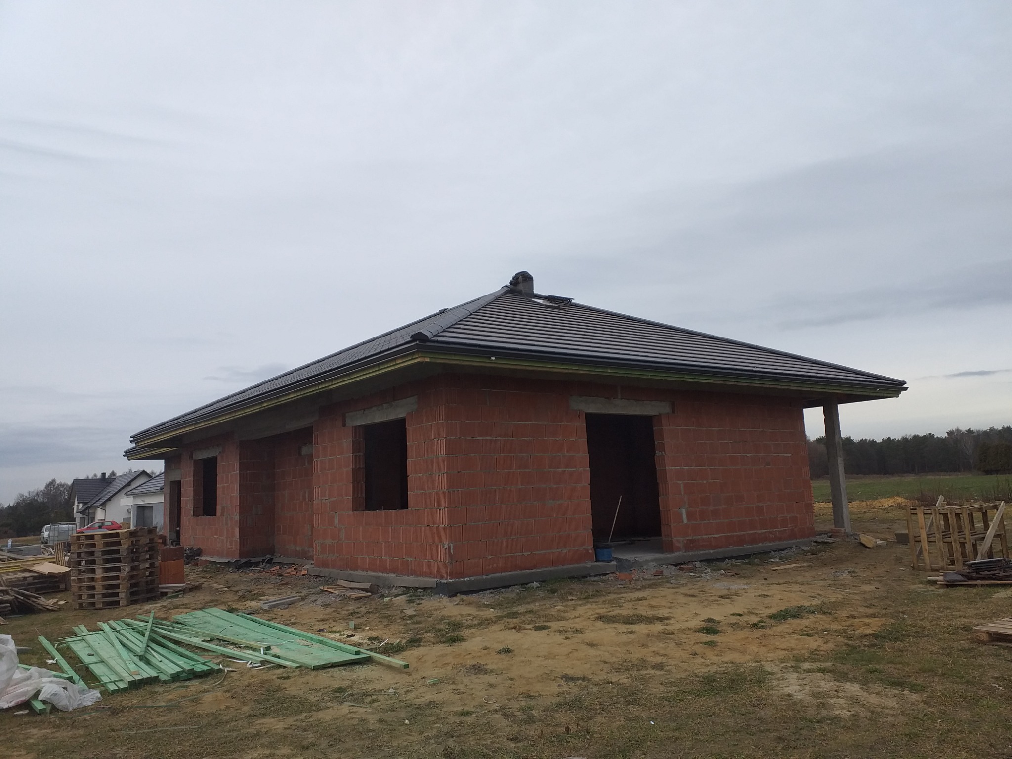 Dom jednorodzinny w trakcie budowy: murowane ściany z czerwonej cegły, zamontowany ciemny dach z rynnami, widoczne palety i materiały budowlane na zewnątrz, pochmurne niebo.