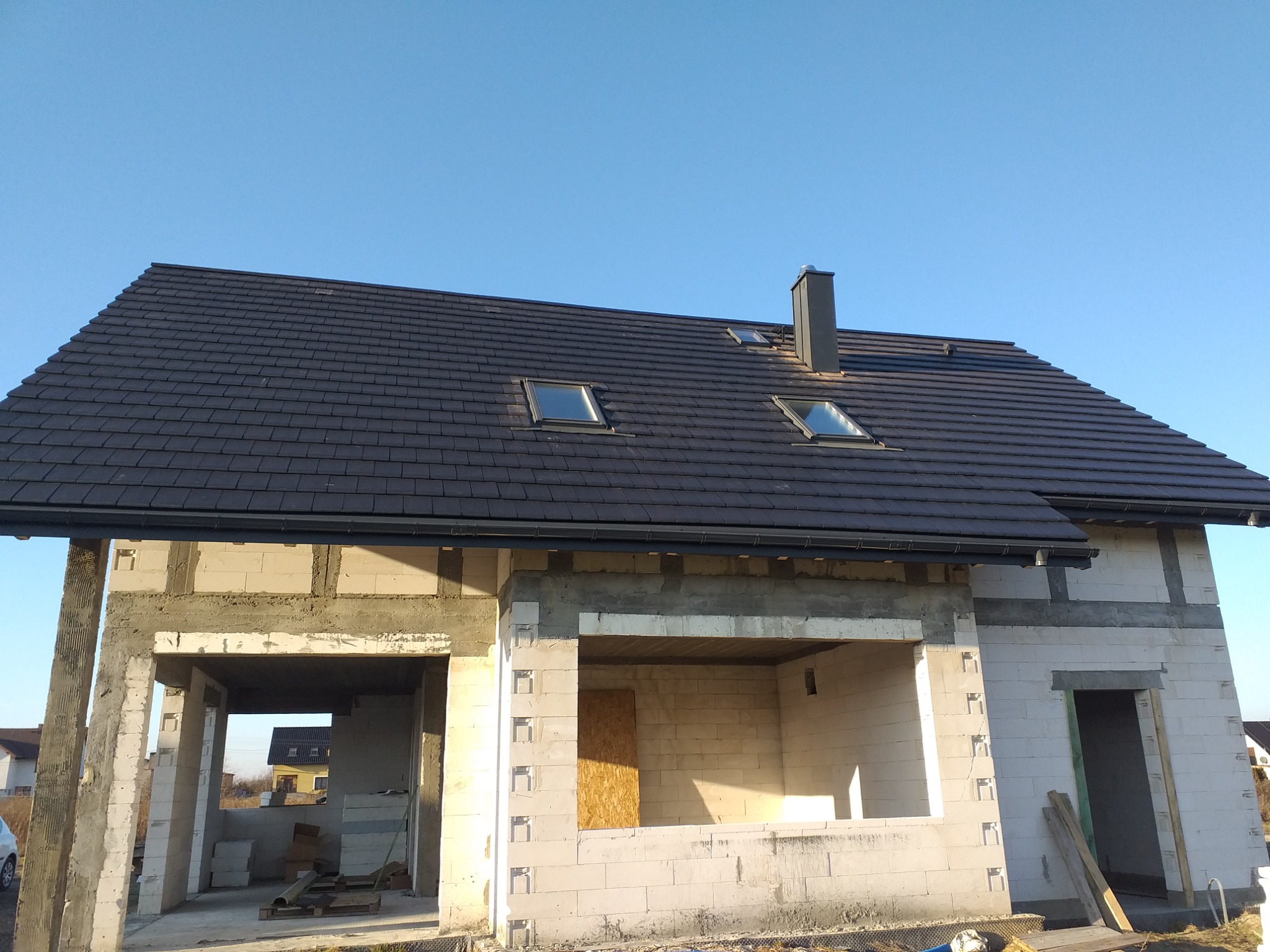 Dom w trakcie budowy z ciemną dachówką, widoczne okna dachowe i komin na tle błękitnego nieba, ściany z betonu komórkowego w stanie surowym.