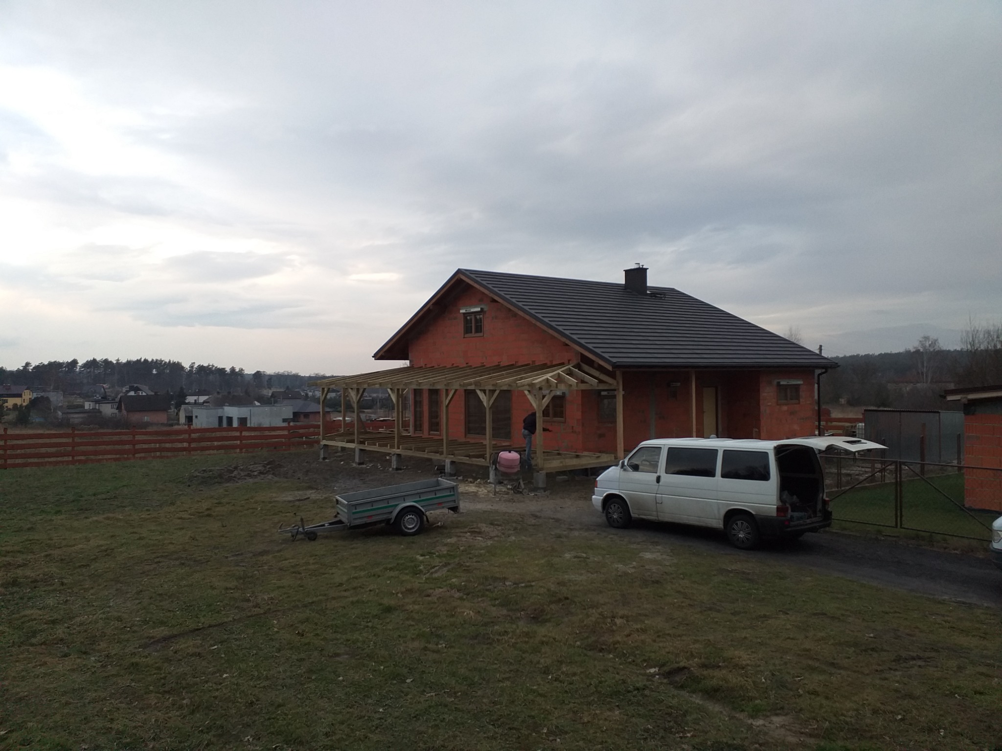 Dom w trakcie budowy z ciemnoszarą dachówką, widoczna drewniana konstrukcja zadaszenia tarasu, przyczepka i biały van z otwartymi drzwiami bagażnika.