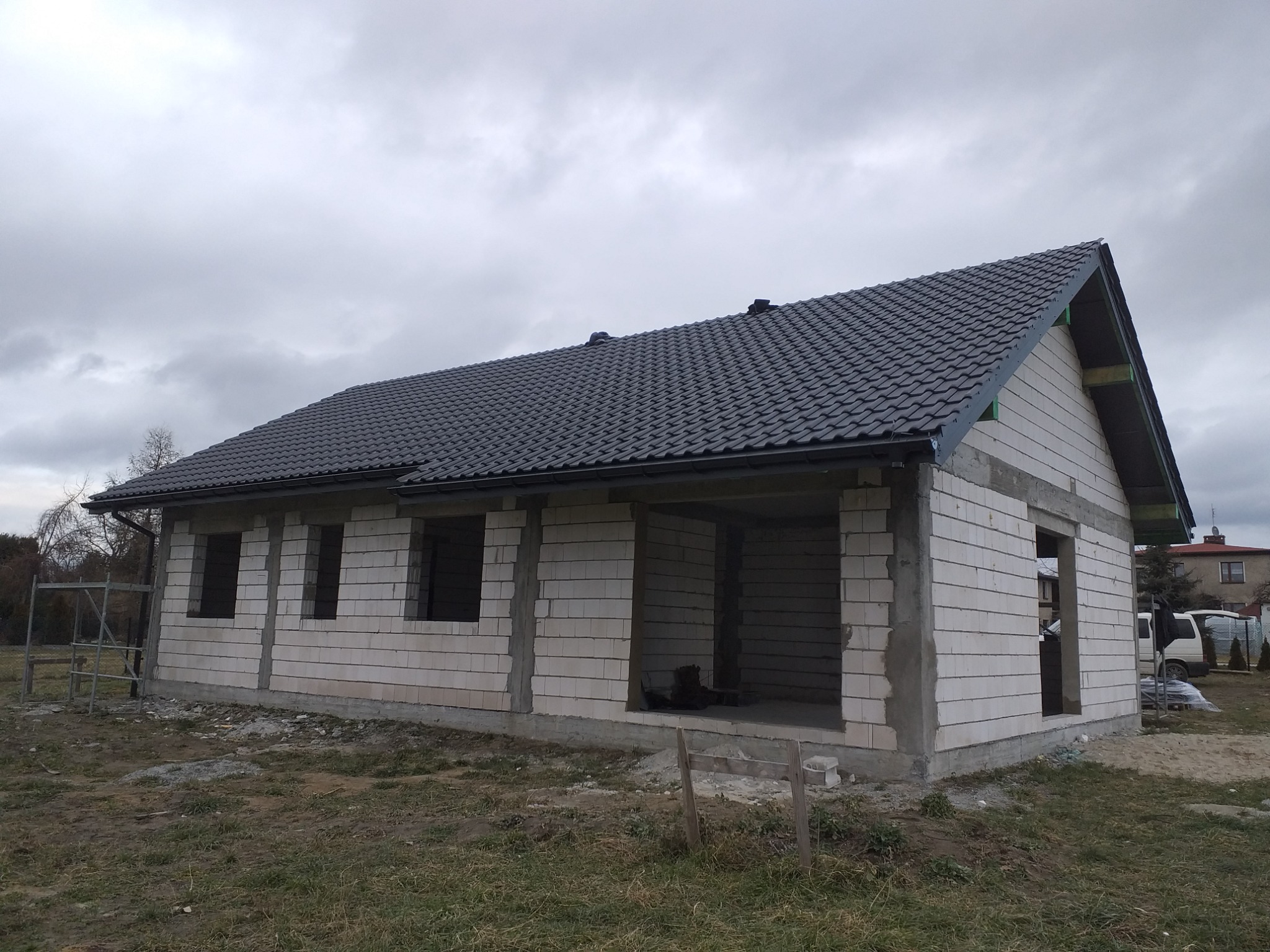 Surowy stan domu jednorodzinnego z ciemną dachówką ceramiczną, widoczne niezadaszone otwory okienne i drzwiowe, szare niebo w tle.