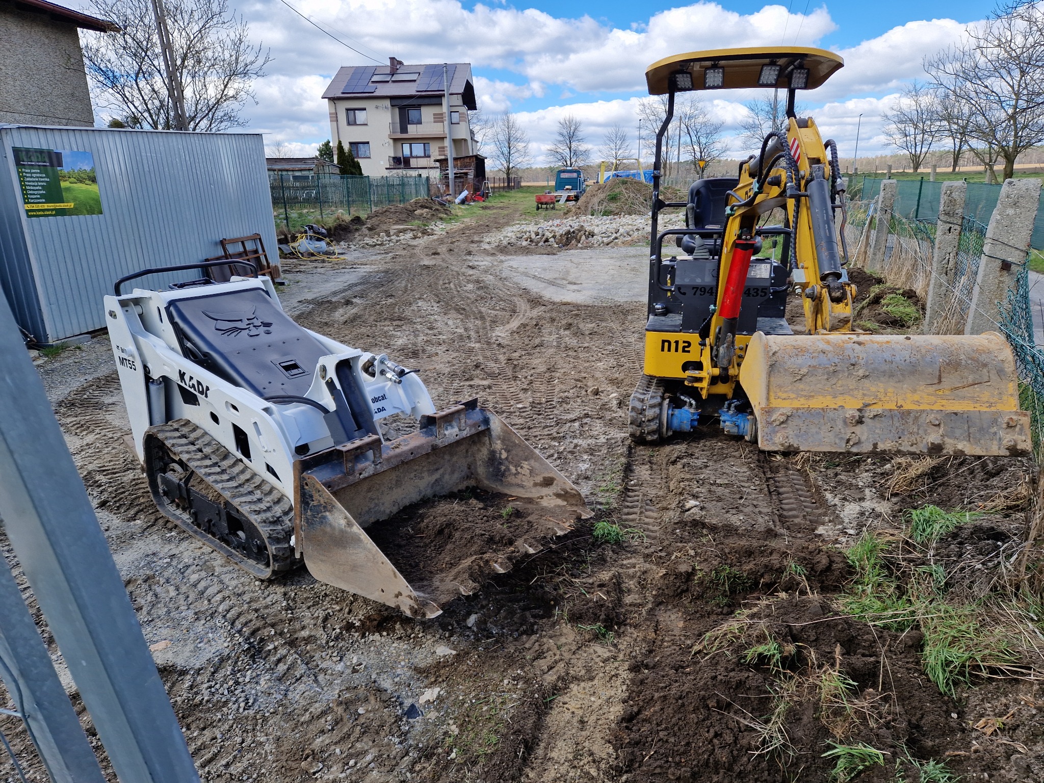 Dwa urządzenia budowlane: biała miniładowarka gąsienicowa i żółta minikoparka na placu budowy z widocznym budynkiem mieszkalnym w tle, ślady opon w błocie.