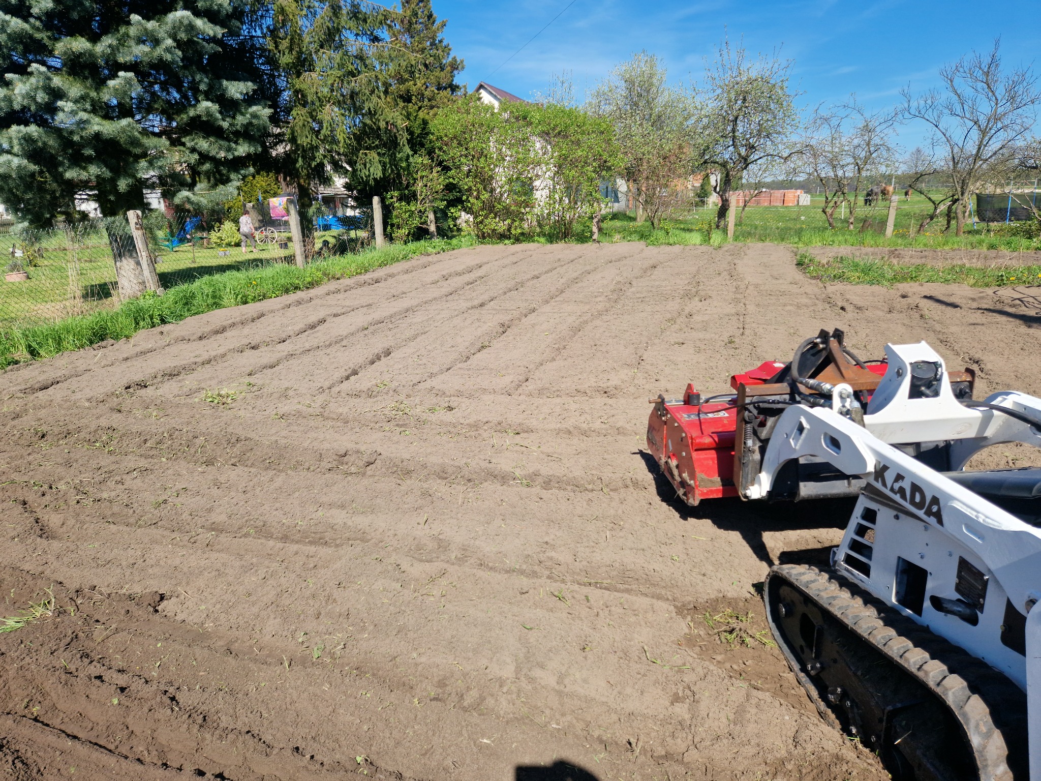 Przygotowane pole pod uprawę z użyciem glebogryzarki KADA na gąsienicach, widoczne rzędy spulchnionej ziemi, w tle zieleń drzew i zabudowania gospodarcze.