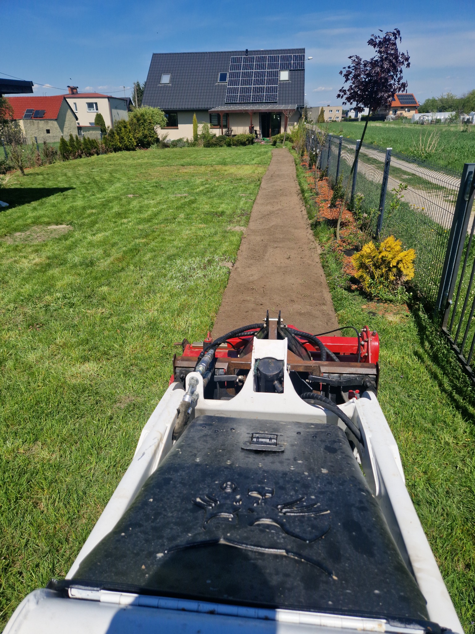 Widok z perspektywy operatora na miniładowarkę z frezarką glebową wyrównującą pas terenu pod przyszły trawnik w ogrodzie, w tle dom z panelami słonecznymi.