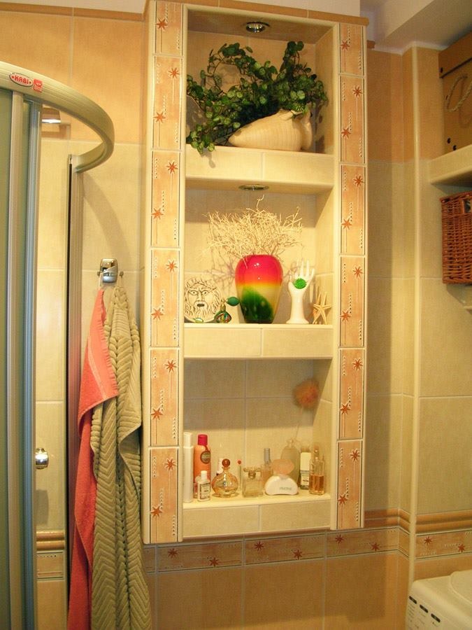 Narożna półka w łazience, wyłożona beżowymi płytkami z motywem gwiazdek, eksponująca dekoracje i kosmetyki na trzech poziomach, z oświetleniem punktowym na górze i widoczną kabiną prysznicową po...