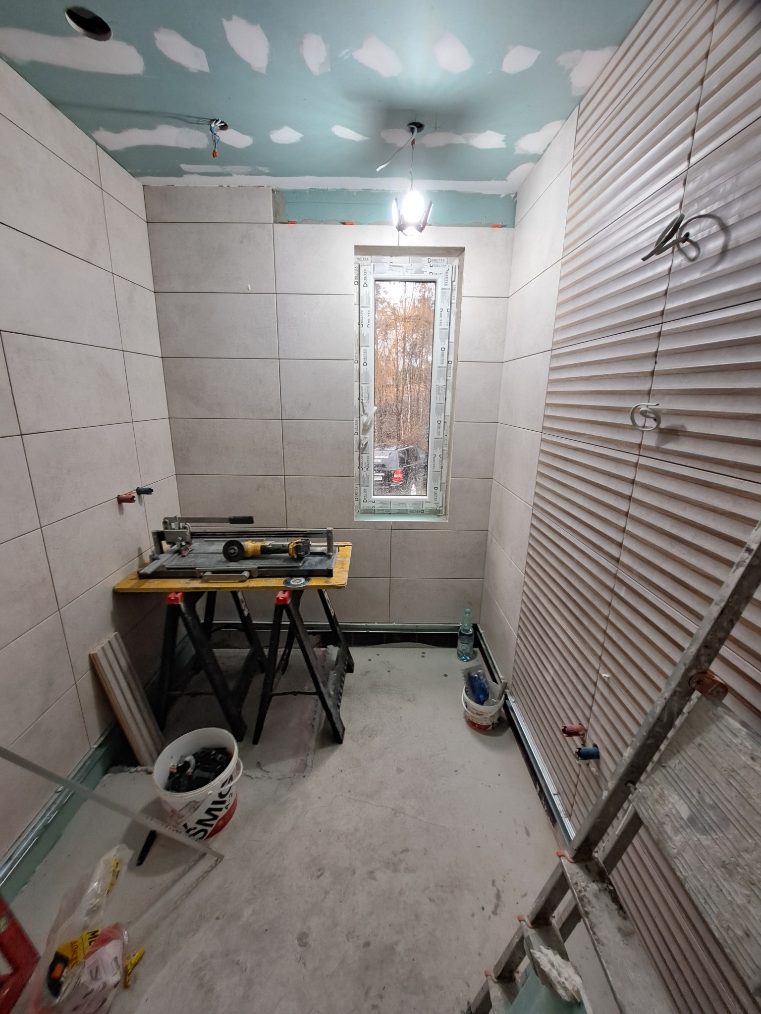 Łazienka w trakcie remontu: ściany wyłożone jasnymi płytkami, jedna ściana z dekoracyjnymi panelami, okno z widokiem na drzewa, narzędzia na stole i drabina. Sufit częściowo pokryty gładzią.