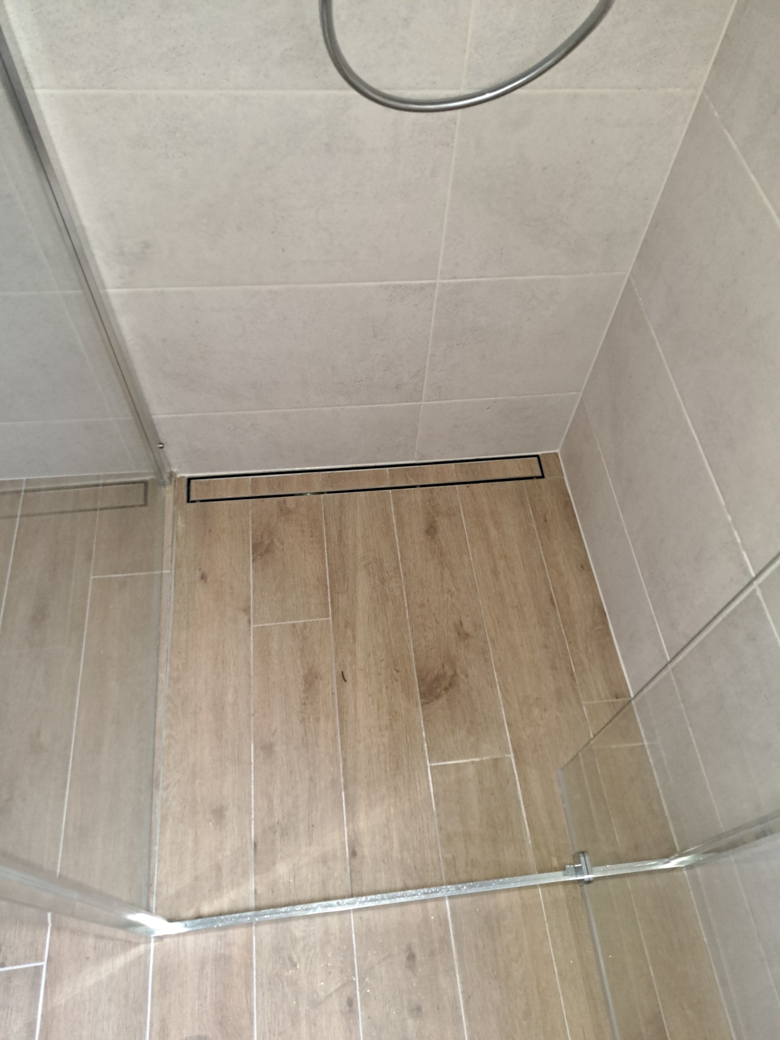 Kabina prysznicowa z odpływem liniowym w podłodze wyłożonej płytkami imitującymi drewno, ściany wyłożone jasnymi płytkami ceramicznymi.