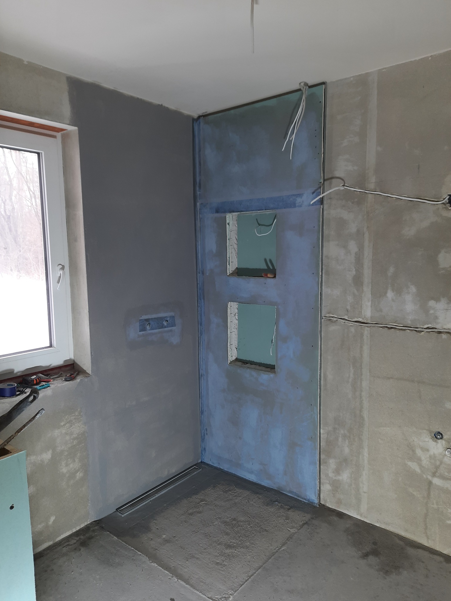Wnętrze łazienki w trakcie remontu: zamontowany odpływ liniowy w posadzce, ściany przygotowane pod płytki, widoczne instalacje elektryczne i otwory rewizyjne w obudowie. Częściowo położona gładź.
