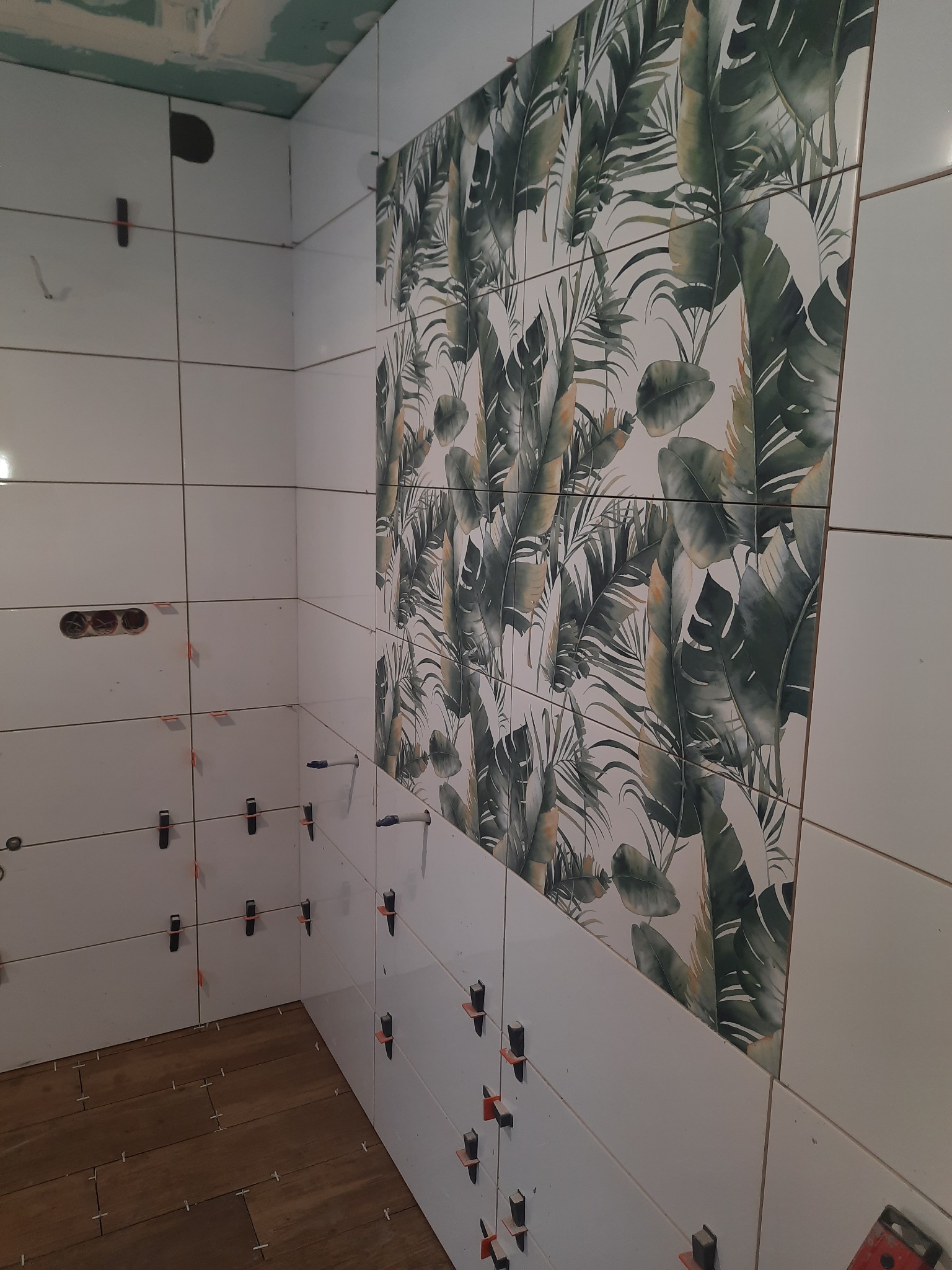 Ściana w trakcie układania białych płytek z motywem liści tropikalnych, widoczne krzyżyki dystansowe i otwory na gniazdka elektryczne, podłoga częściowo wyłożona panelami imitującymi drewno.
