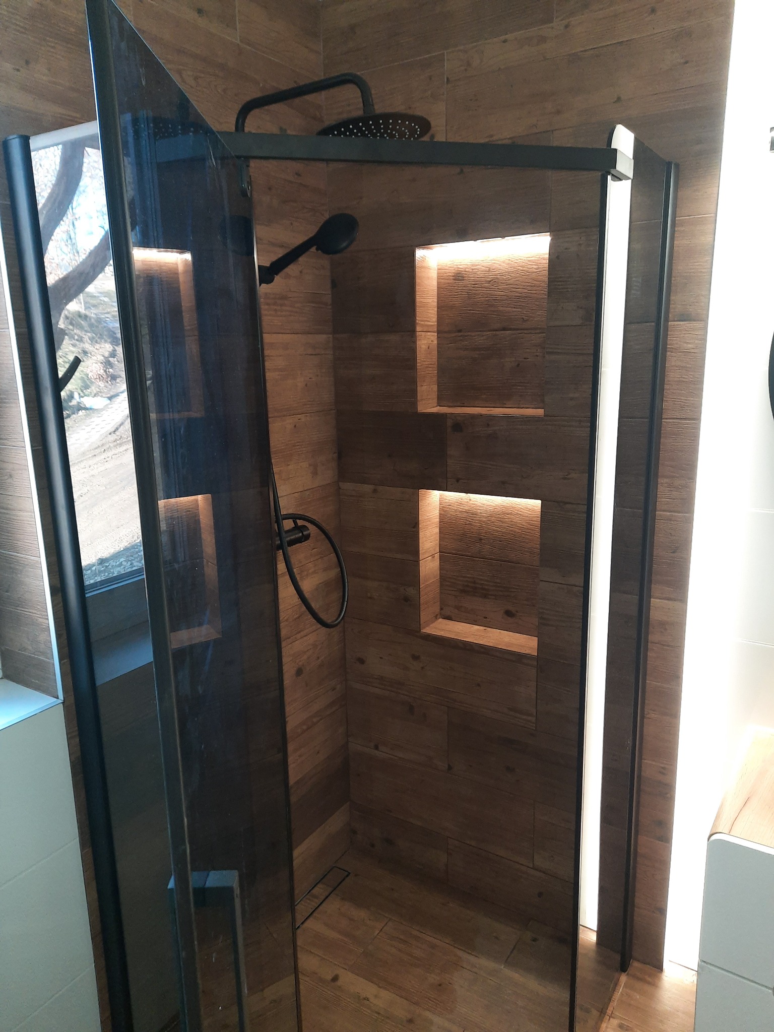 Kabina prysznicowa z czarnymi profilami i deszczownicą, drewnopodobne płytki na ścianach i podłodze, w ścianie wnęki z oświetleniem.