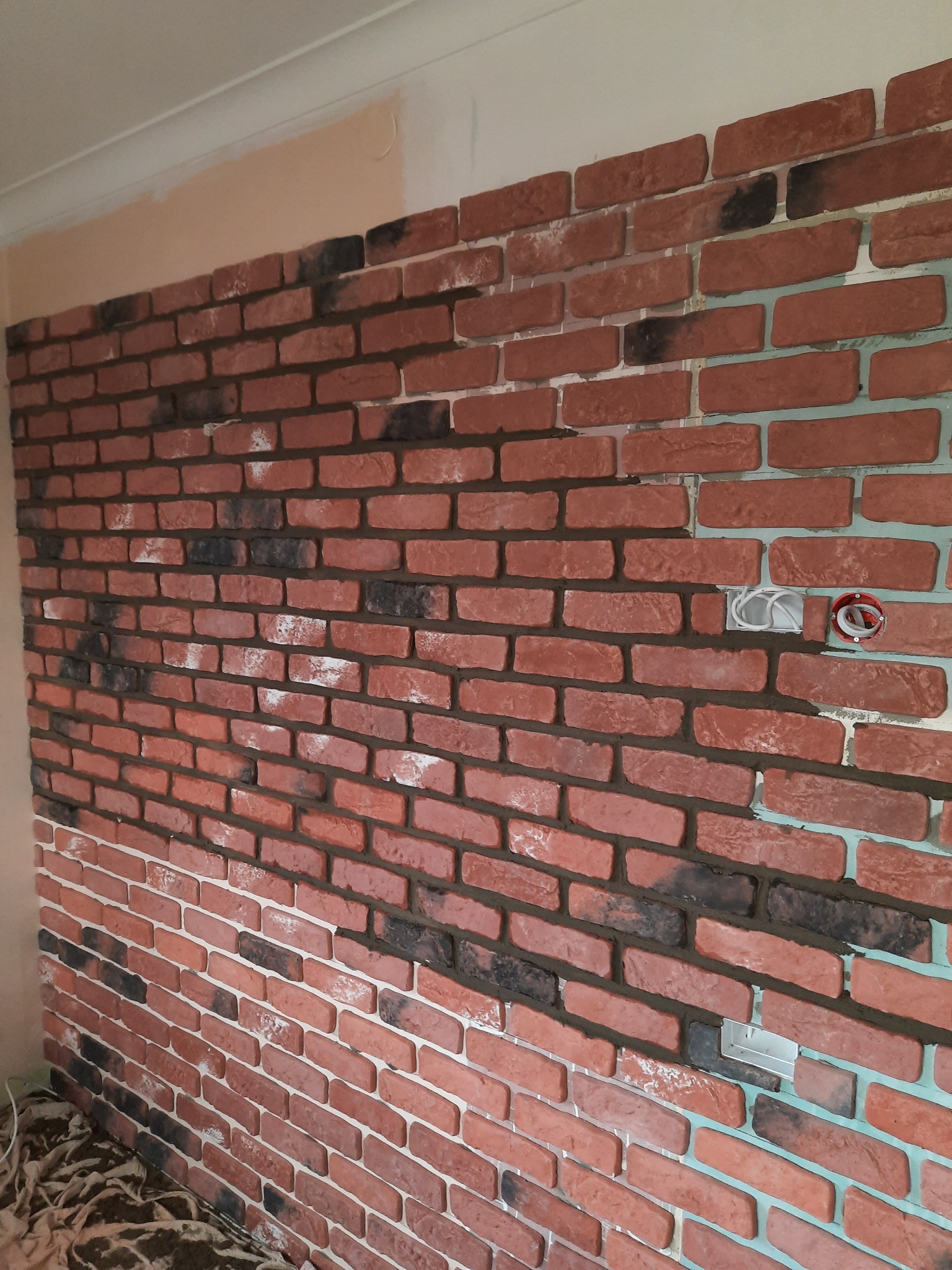 Ściana wykończona czerwoną cegłą dekoracyjną z elementami czarnej cegły, widoczne puszki elektryczne i fragmenty ściany przed wykończeniem.