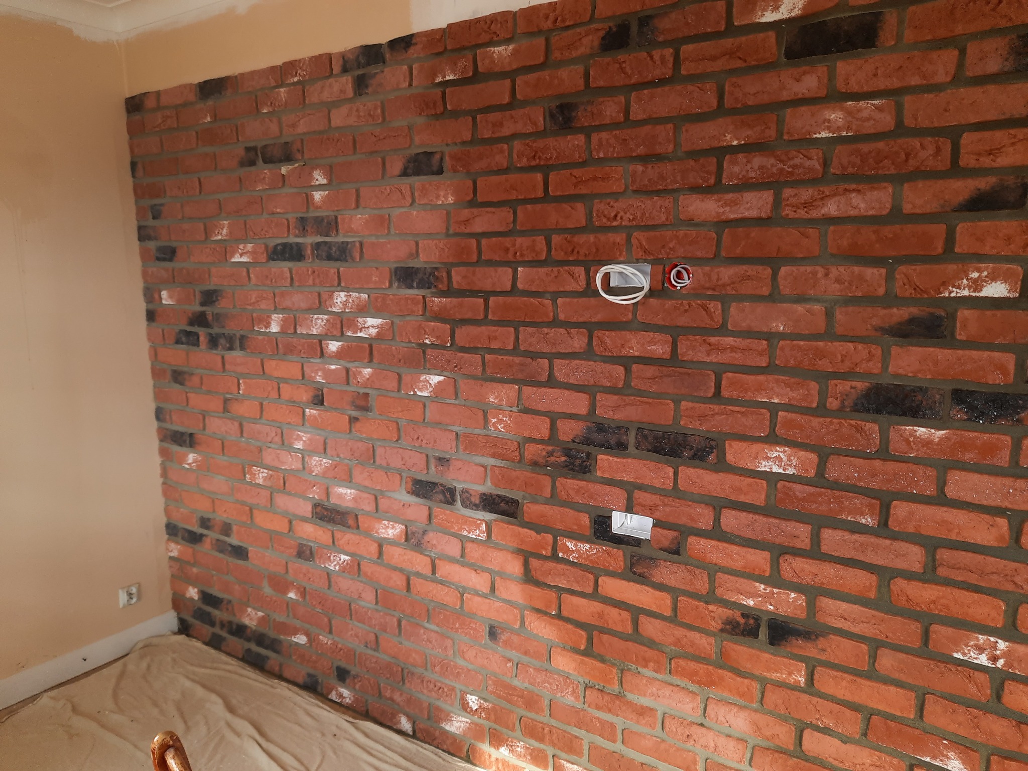 Ściana wyłożona dekoracyjną cegłą w odcieniach czerwieni i czerni, z widocznymi gniazdkami elektrycznymi i kablem, zabezpieczona folią malarską przy podłodze.