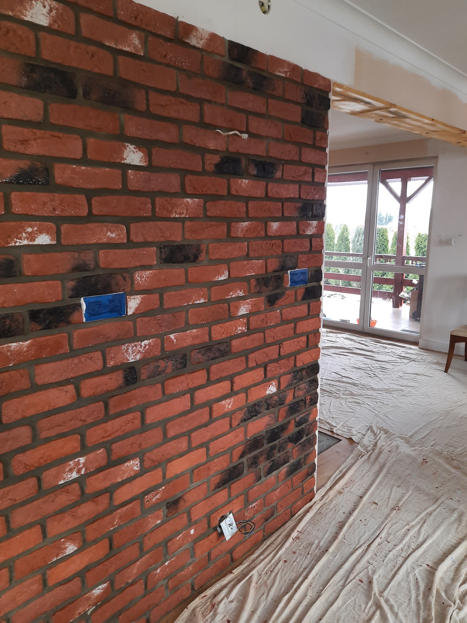 Ściana wyłożona czerwoną cegłą klinkierową, z widocznymi śladami białej farby i dwoma niebieskimi elementami maskującymi, podłoga zabezpieczona folią malarską, widok na balkon w tle.