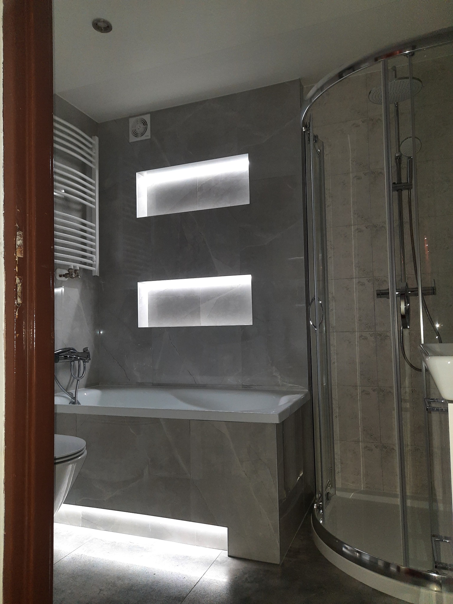 Nowoczesna łazienka wykończona szarymi płytkami imitującymi marmur, z podświetlanymi wnękami nad wanną i kabiną prysznicową z zaokrąglonymi drzwiami, widoczny fragment toalety i grzejnika...
