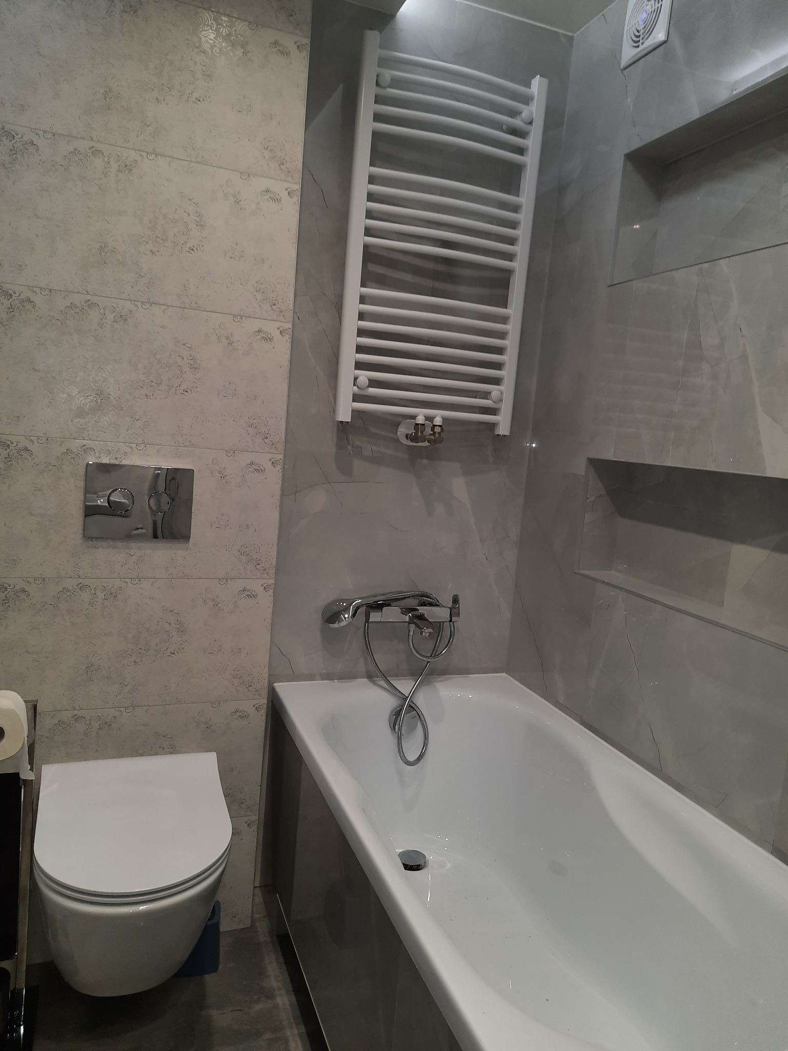 Nowoczesna łazienka w szarościach z wiszącą toaletą, wanną i grzejnikiem drabinkowym; płytki z delikatnym wzorem na ścianach.