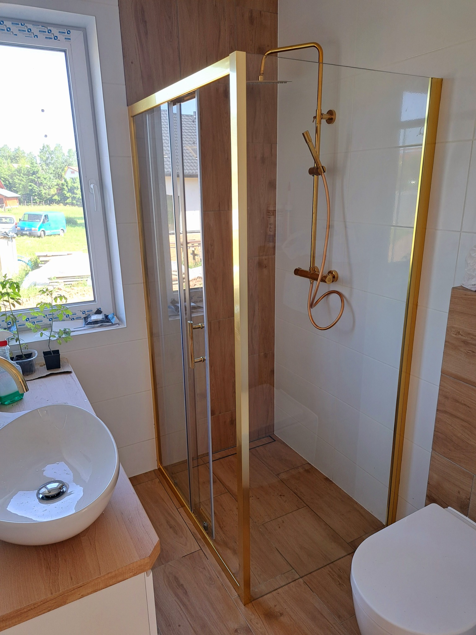 Nowoczesna łazienka z kabiną prysznicową w złotej ramie, armaturą w kolorze miedzi, umywalką nablatową i drewnopodobnymi płytkami na podłodze i ścianie.