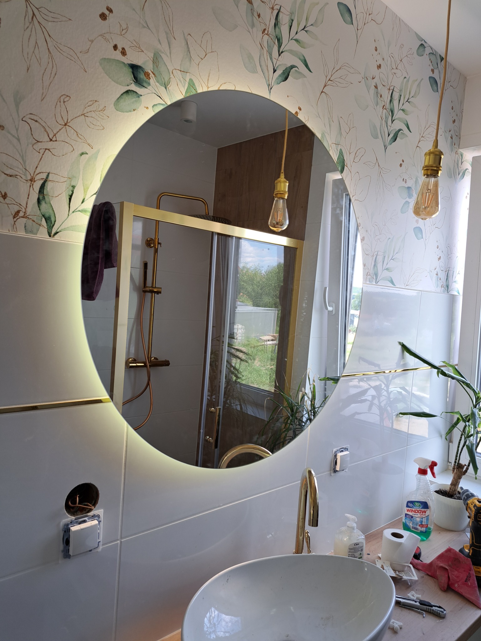 Okrągłe lustro z podświetleniem LED odbija złotą kabinę prysznicową w łazience z motywem roślinnym na tapecie, widoczny fragment umywalki, baterii, gniazdka elektrycznego i akcesoriów łazienkowych.