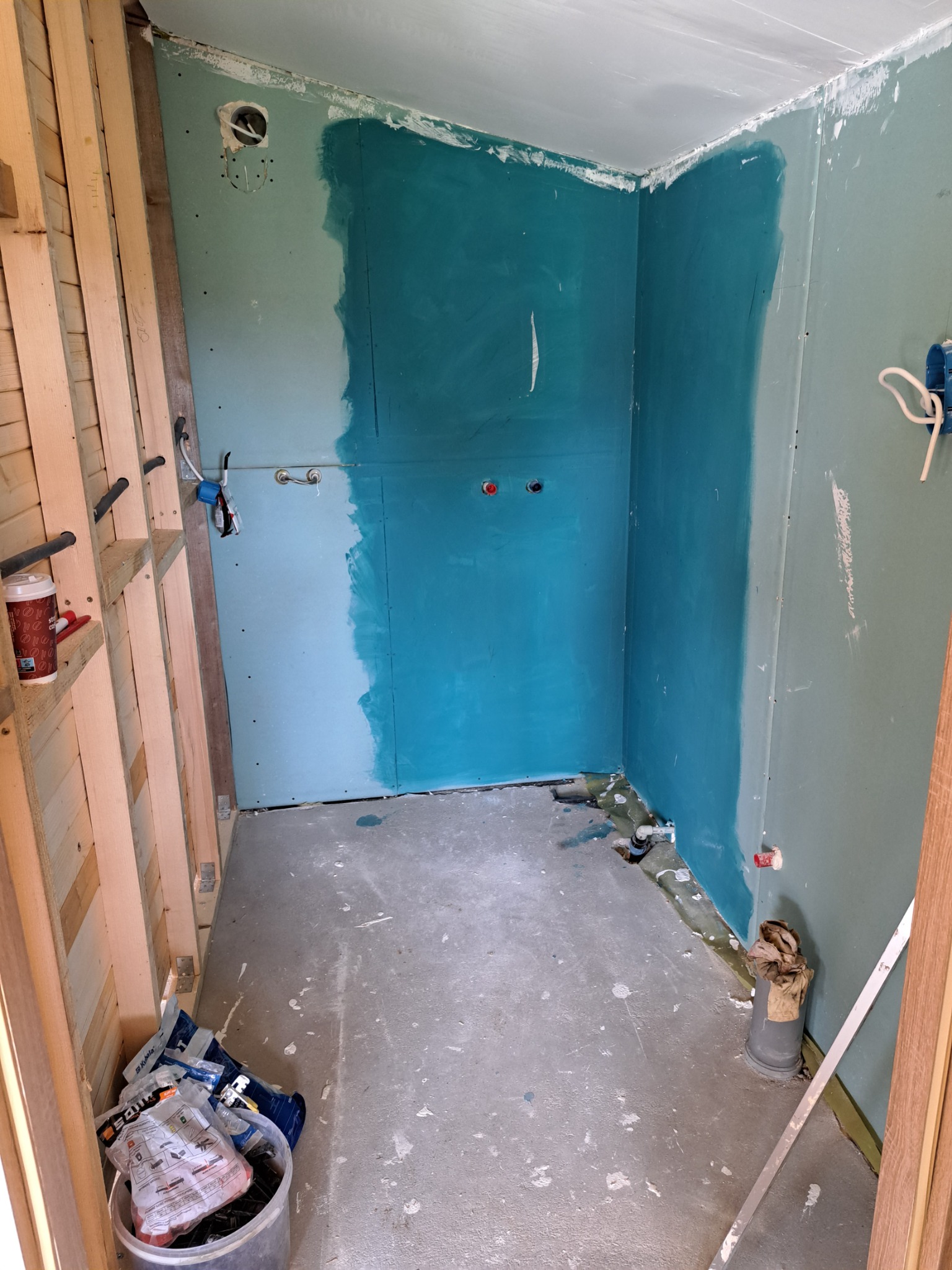 Wnętrze łazienki w trakcie remontu: częściowo pomalowane na turkusowo ściany z płyt kartonowo-gipsowych, widoczne instalacje, konstrukcja drewniana, narzędzia i materiały budowlane na podłodze.