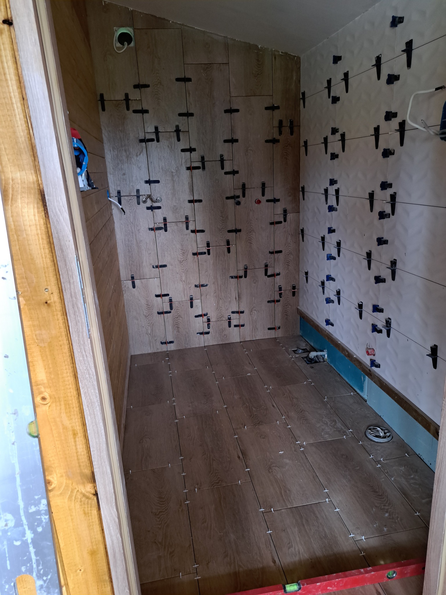 Wnętrze łazienki w trakcie układania płytek imitujących drewno na podłodze i ścianach, z widocznymi klipsami i klinami poziomującymi, oraz poziomnicą na podłodze.