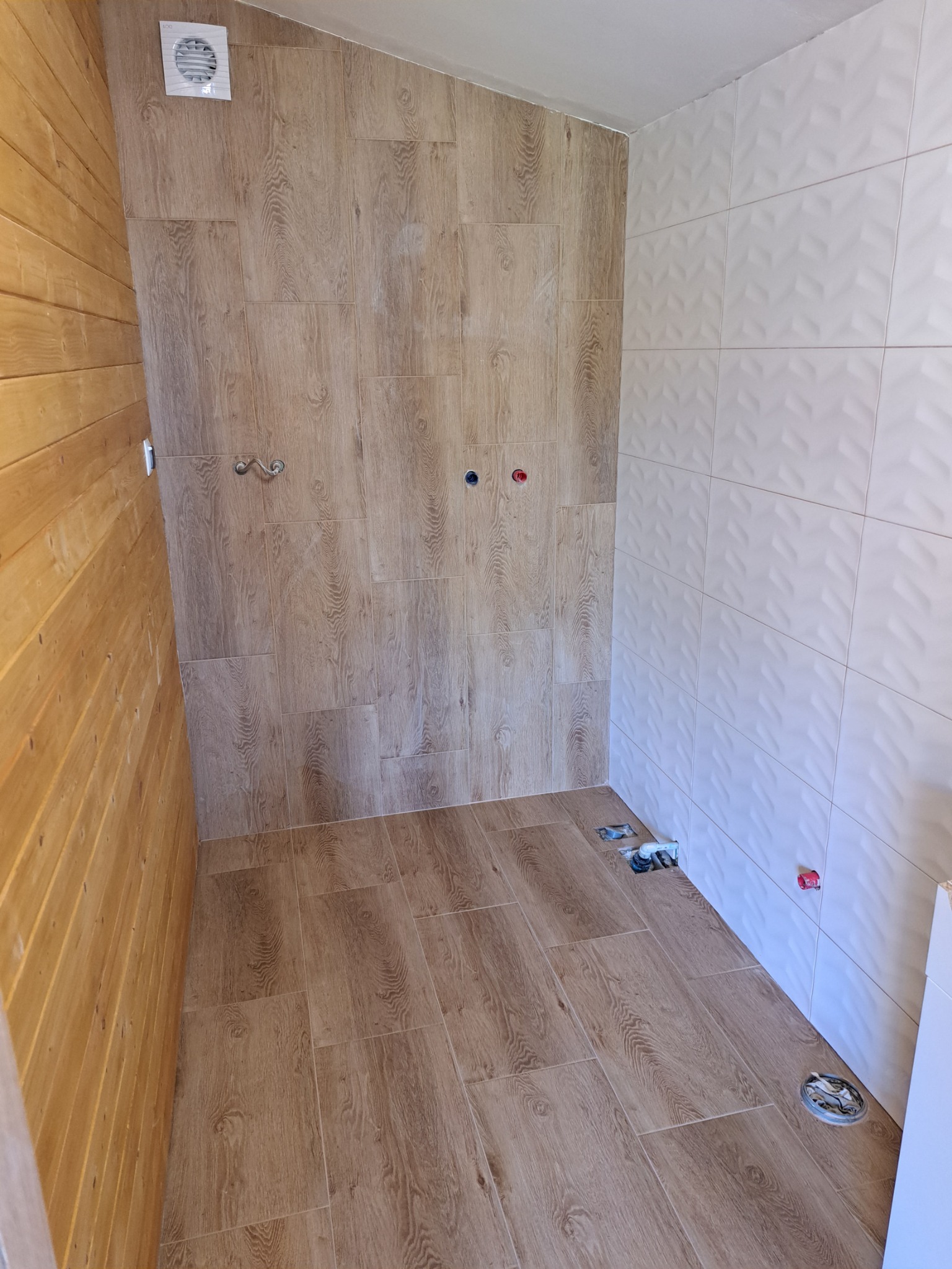 Wnętrze łazienki w trakcie remontu, z wyłożonymi płytkami imitującymi drewno na podłodze i części ścian, oraz białymi płytkami 3D na pozostałej części ścian. Widoczne wystające rury i otwory...