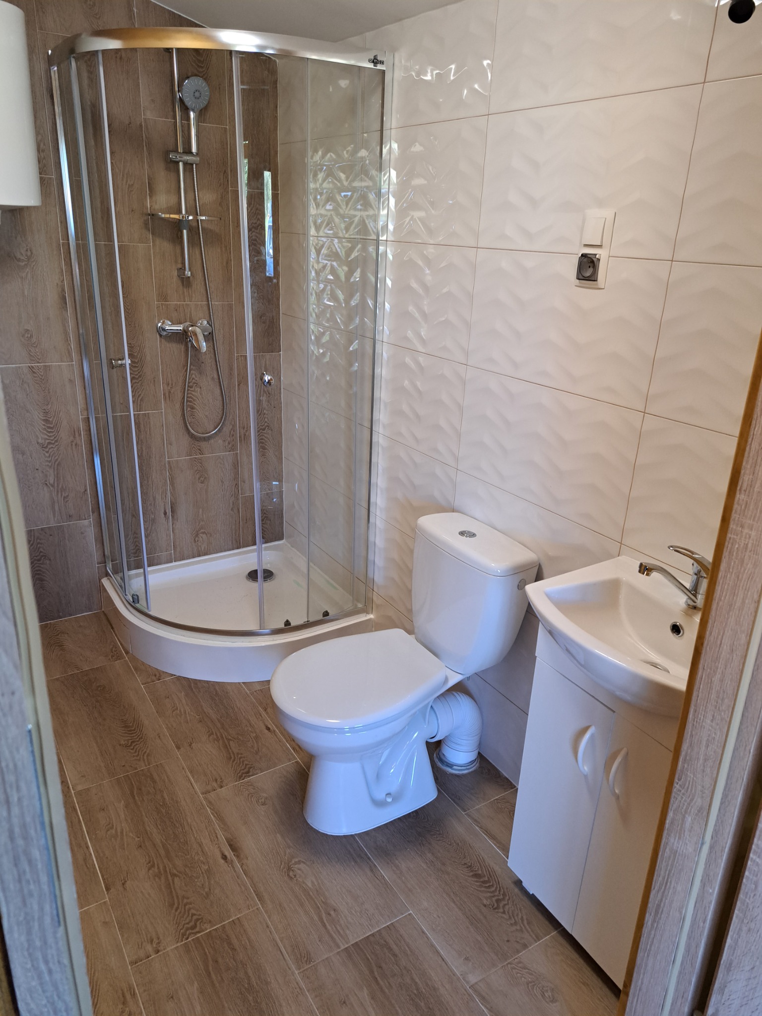 Nowoczesna łazienka po remoncie: kabina prysznicowa narożna z chromowaną armaturą, biała ceramika sanitarna (toaleta i umywalka z szafką), płytki imitujące drewno na podłodze oraz białe płytki...