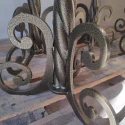 Metalowa konstrukcja z dekoracyjnymi elementami w kształcie spirali, pokryta farbą o fakturze młotkowej w odcieniach brązu i złota, umieszczona na drewnianych deskach