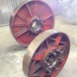 Dwa duże, czerwone koła zamachowe z metalowymi szprychami, leżące na betonowej podłodze w hali przemysłowej, widoczne ślady rdzy i zużycia.