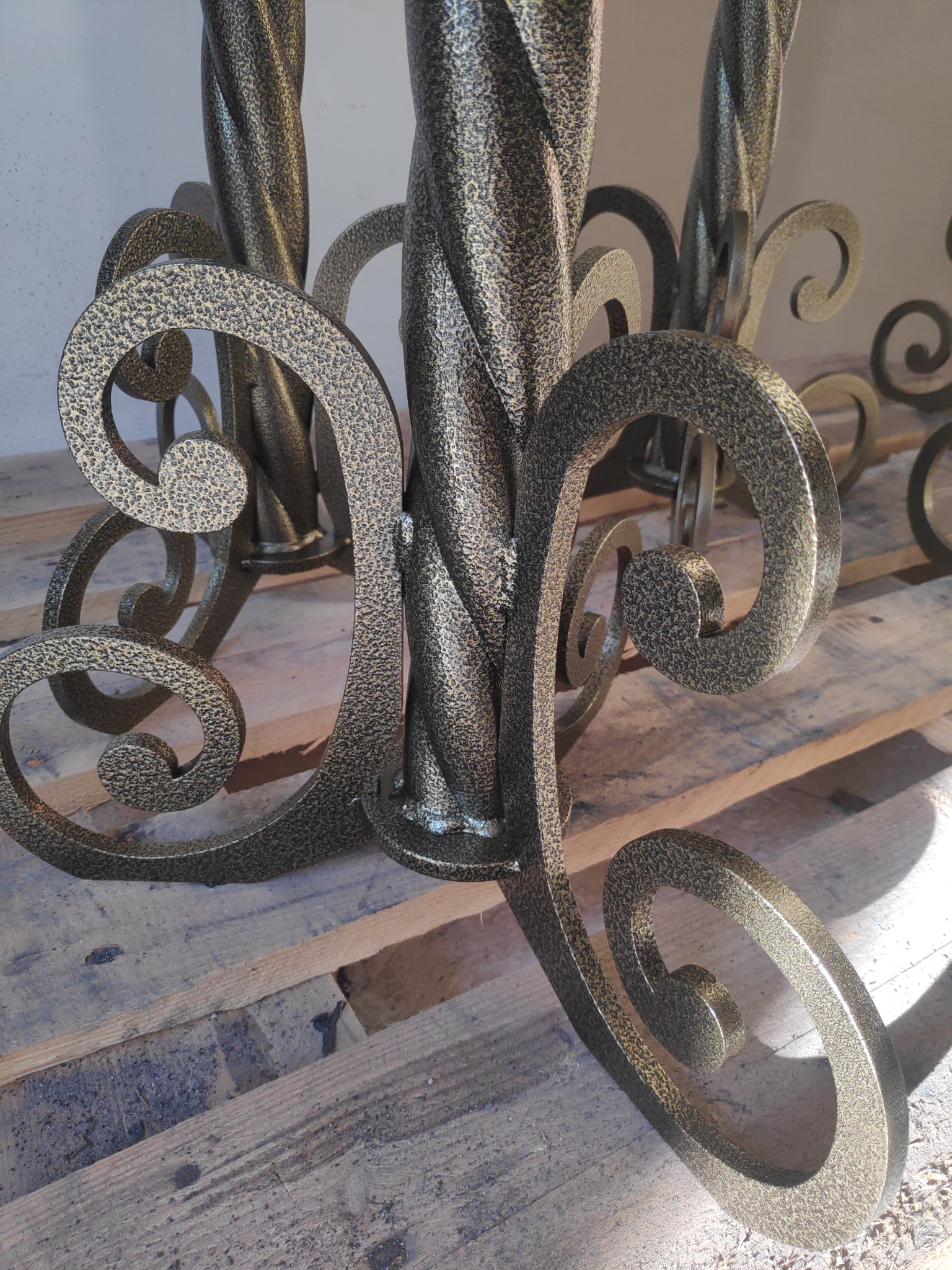 Metalowa konstrukcja z dekoracyjnymi elementami w kształcie spirali, pokryta farbą o fakturze młotkowej w odcieniach brązu i złota, umieszczona na drewnianych deskach