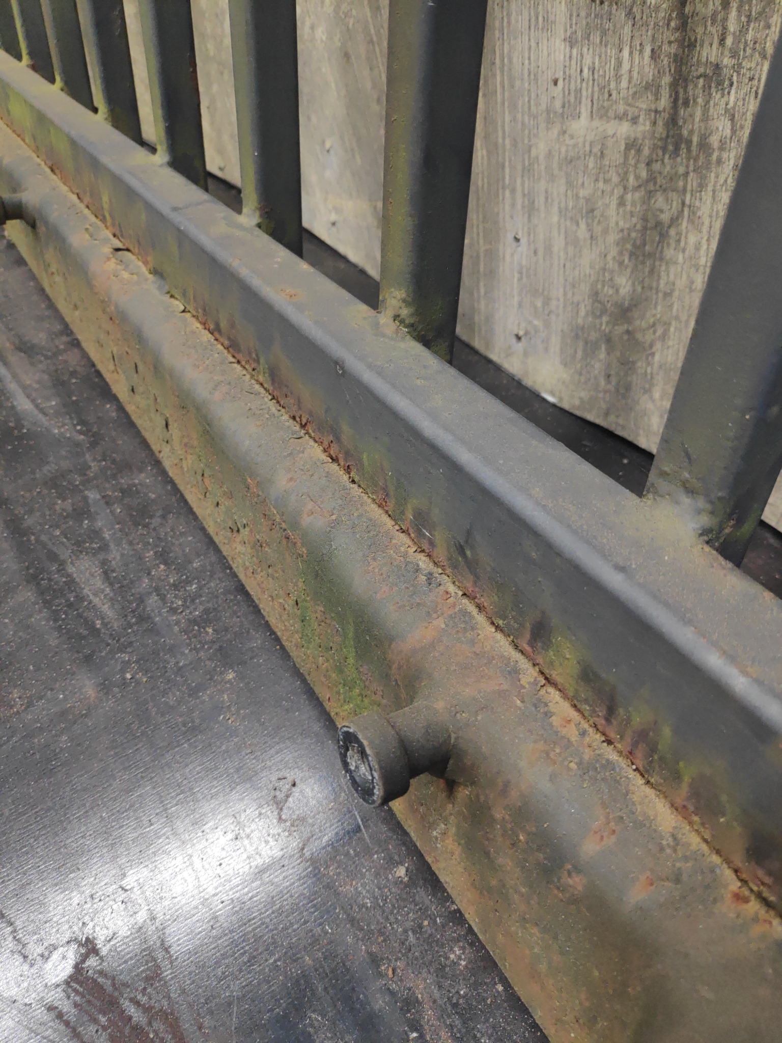 Fragment metalowej konstrukcji z widocznymi śladami rdzy i mchu, pomalowanej na ciemny kolor, z szarym drewnianym tłem.