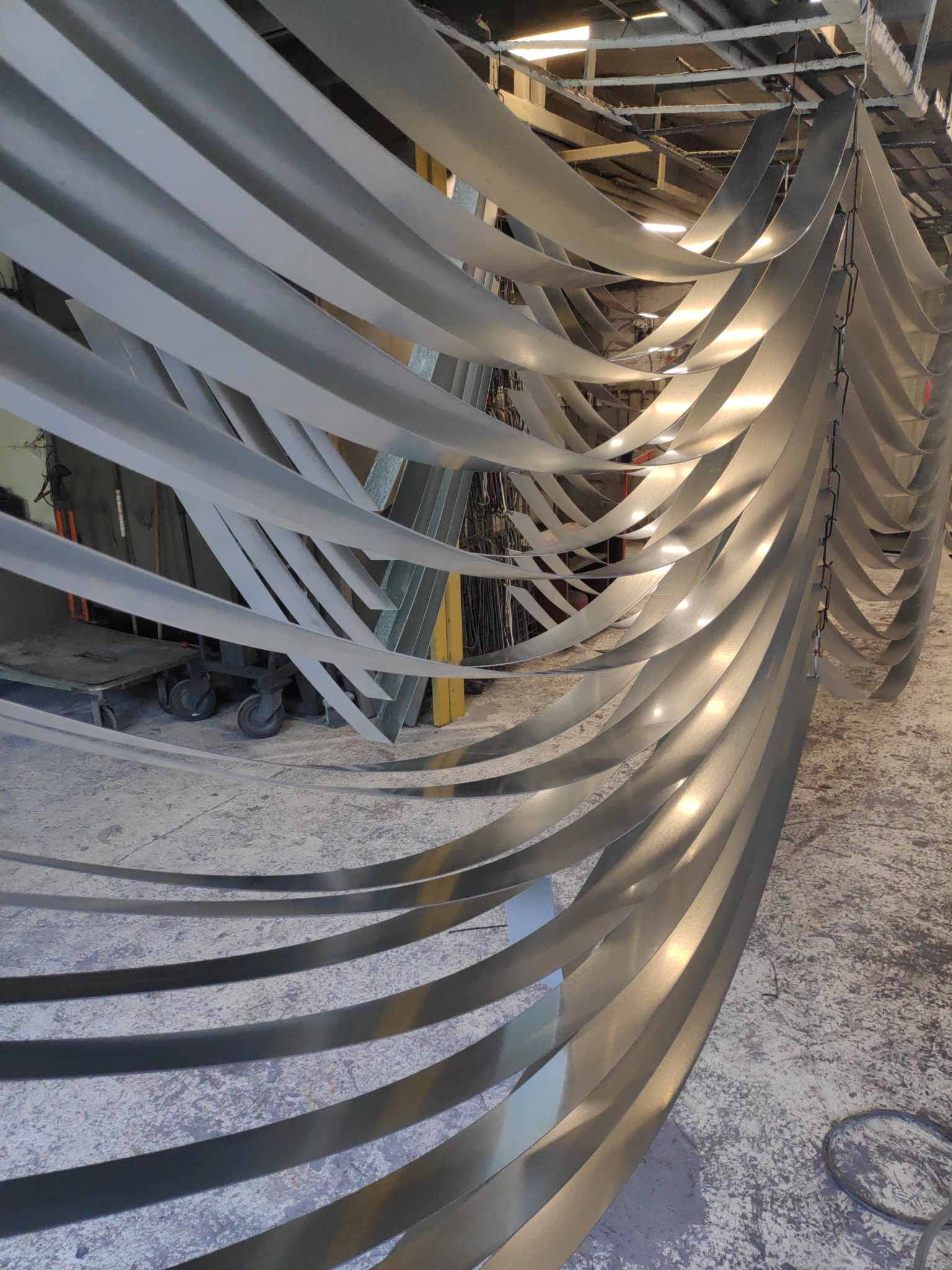 Metalowe, zakrzywione pasy zwisające z sufitu hali, prawdopodobnie po obróbce, z widocznymi refleksami światła.