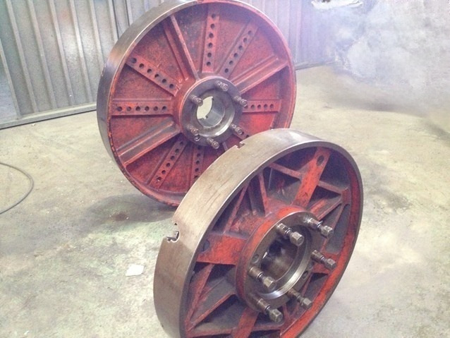 Dwa duże, czerwone koła zamachowe z metalowymi szprychami, leżące na betonowej podłodze w hali przemysłowej, widoczne ślady rdzy i zużycia.