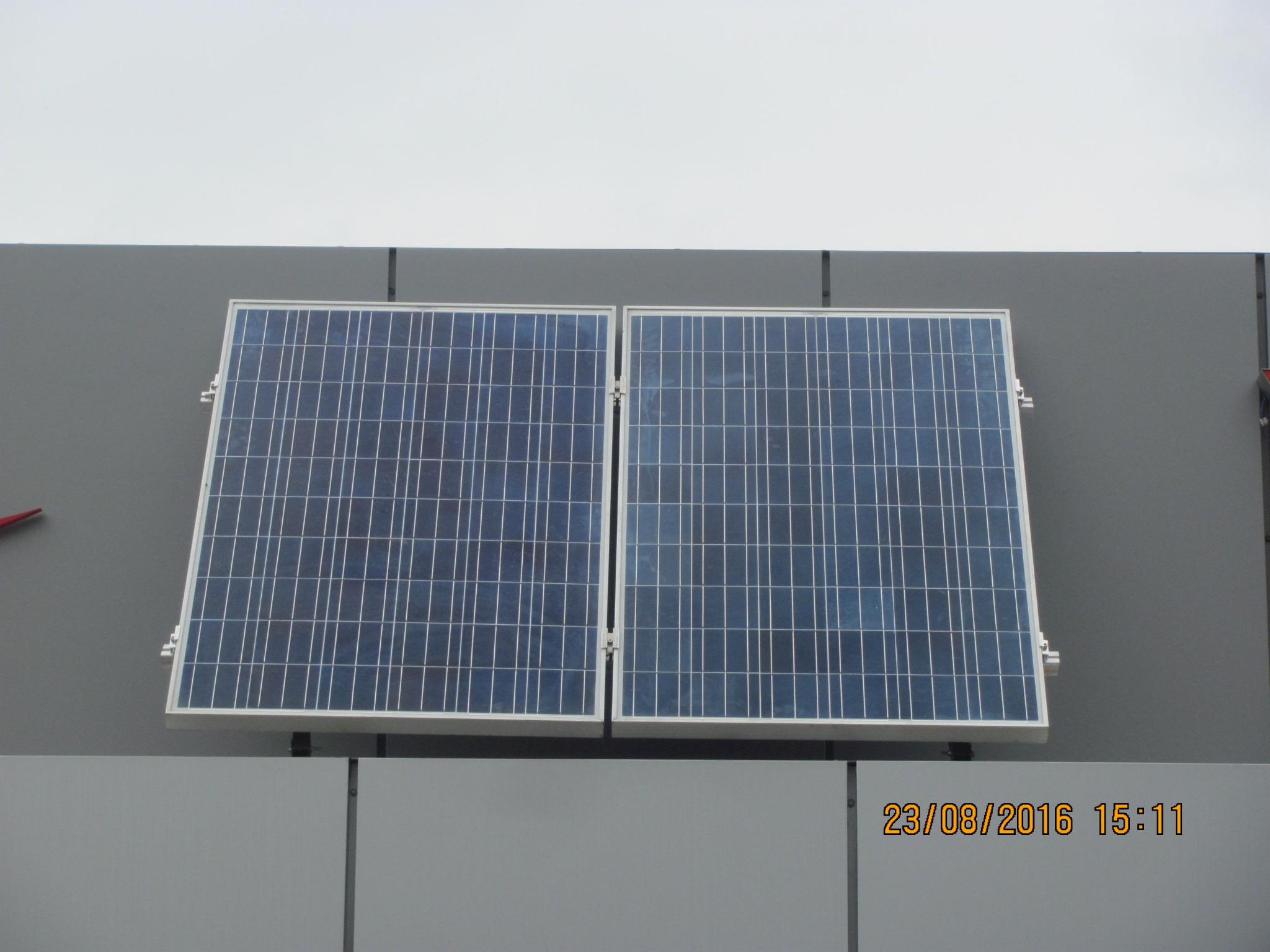 Dwa panele słoneczne zamontowane na szarej ścianie budynku, widoczne mocowania i data wykonania zdjęcia w prawym dolnym rogu.