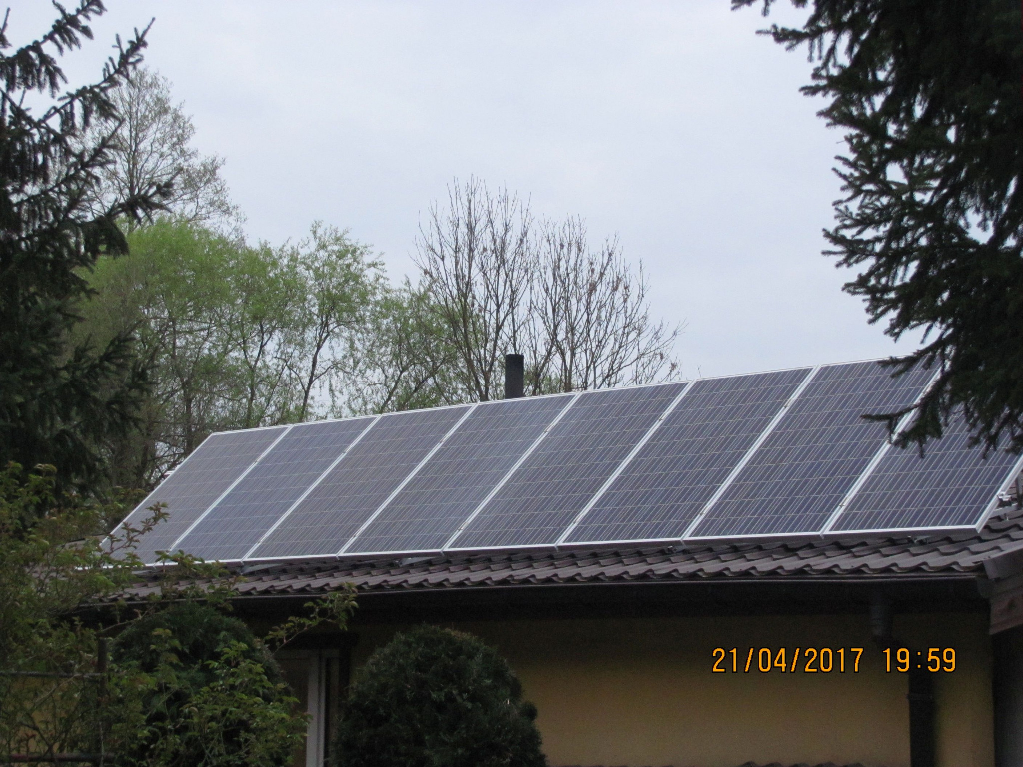 Panele słoneczne zamontowane na brązowym dachu domu jednorodzinnego, widoczne zza drzew i krzewów w pochmurny dzień.