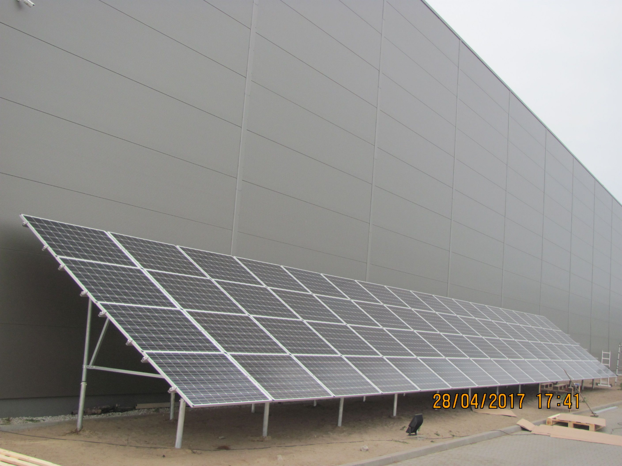 Panel słoneczny zamontowany na ziemi przed szarą ścianą budynku, widoczny pod kątem, z paletami i drabiną w tle.