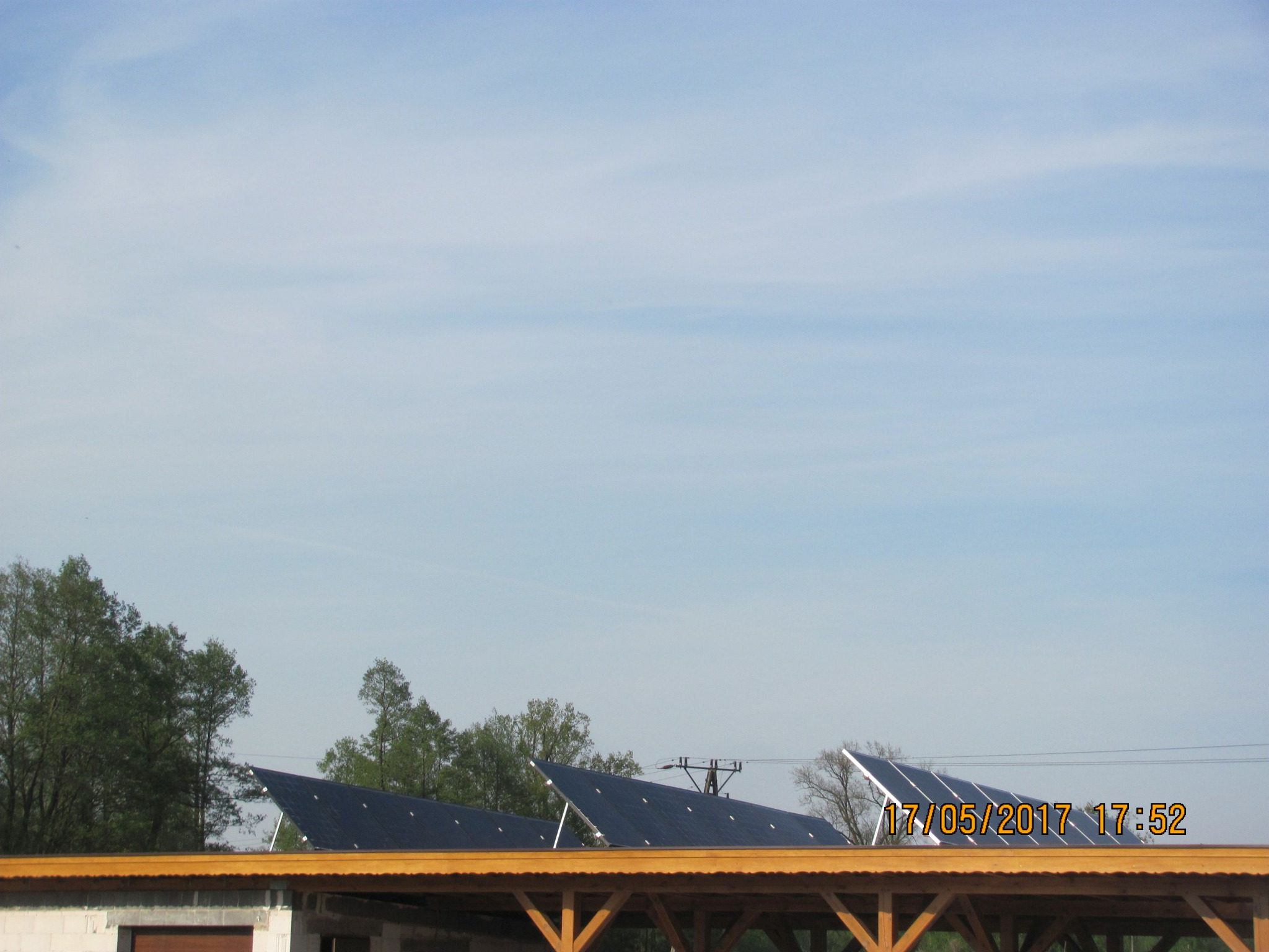 Panele słoneczne zamontowane na drewnianej konstrukcji w słoneczny dzień, widoczne drzewa w tle.