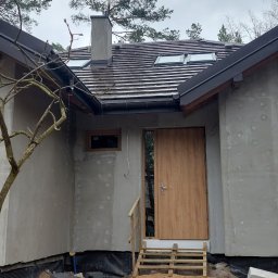 Rem_vm_bud - Surowy stan elewacji domu jednorodzinnego z drewnianymi schodami wejściowymi, widoczny fragment dachu z dachówką i oknami połaciowymi oraz komin.