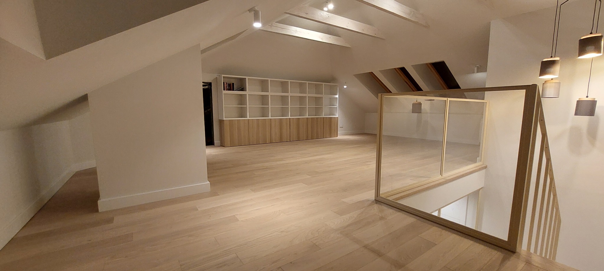 Jasne poddasze po remoncie z drewnianą podłogą, białą zabudową regałową i szklaną balustradą schodów, oświetlone reflektorami i wiszącymi lampami.