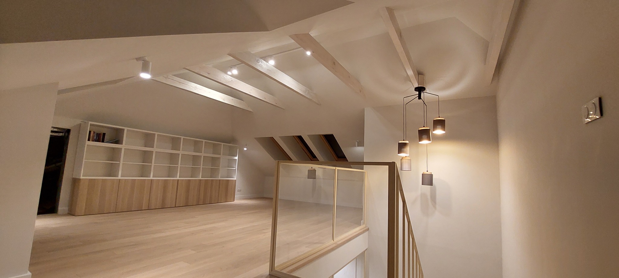 Poddasze po renowacji: drewniana podłoga, zabudowa regałowa z jasnego drewna, drewniane belki stropowe z reflektorami i nowoczesna lampa wisząca na schodach.