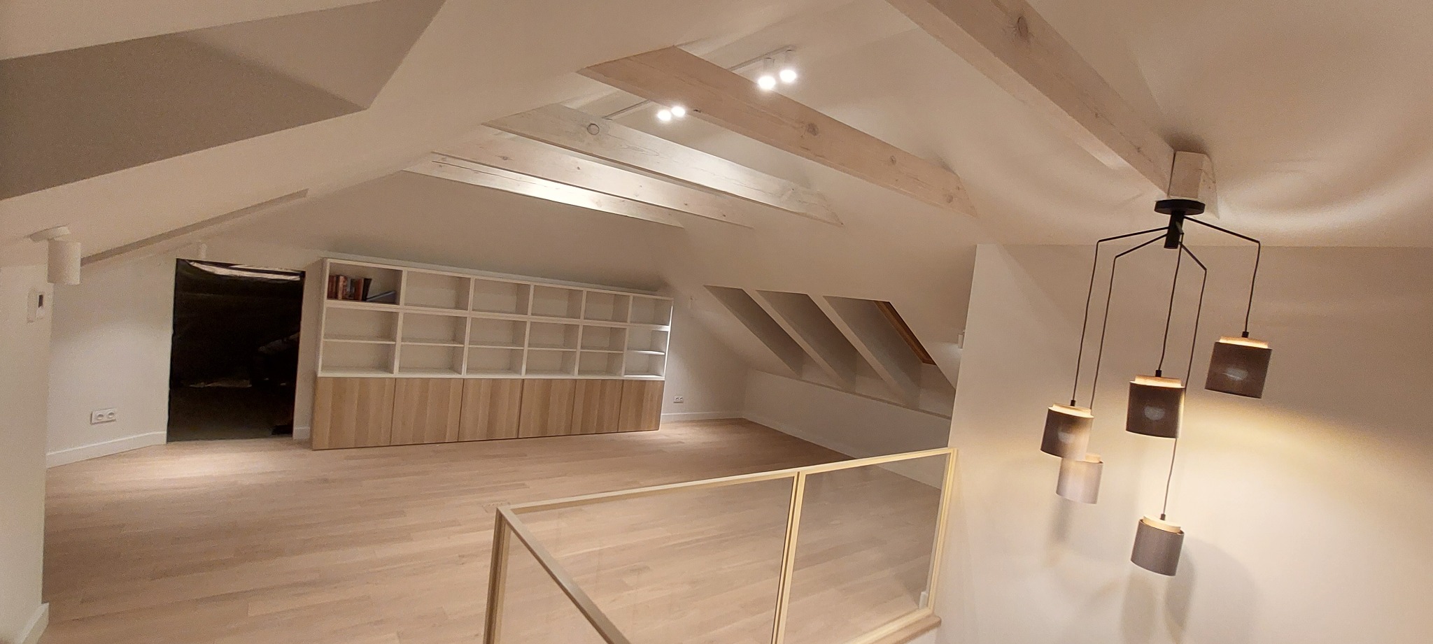Przestronne poddasze z drewnianą podłogą, białe ściany, wbudowane regały z drewnianymi szafkami, widoczne drewniane belki stropowe i nowoczesna lampa wisząca z kilkoma abażurami.