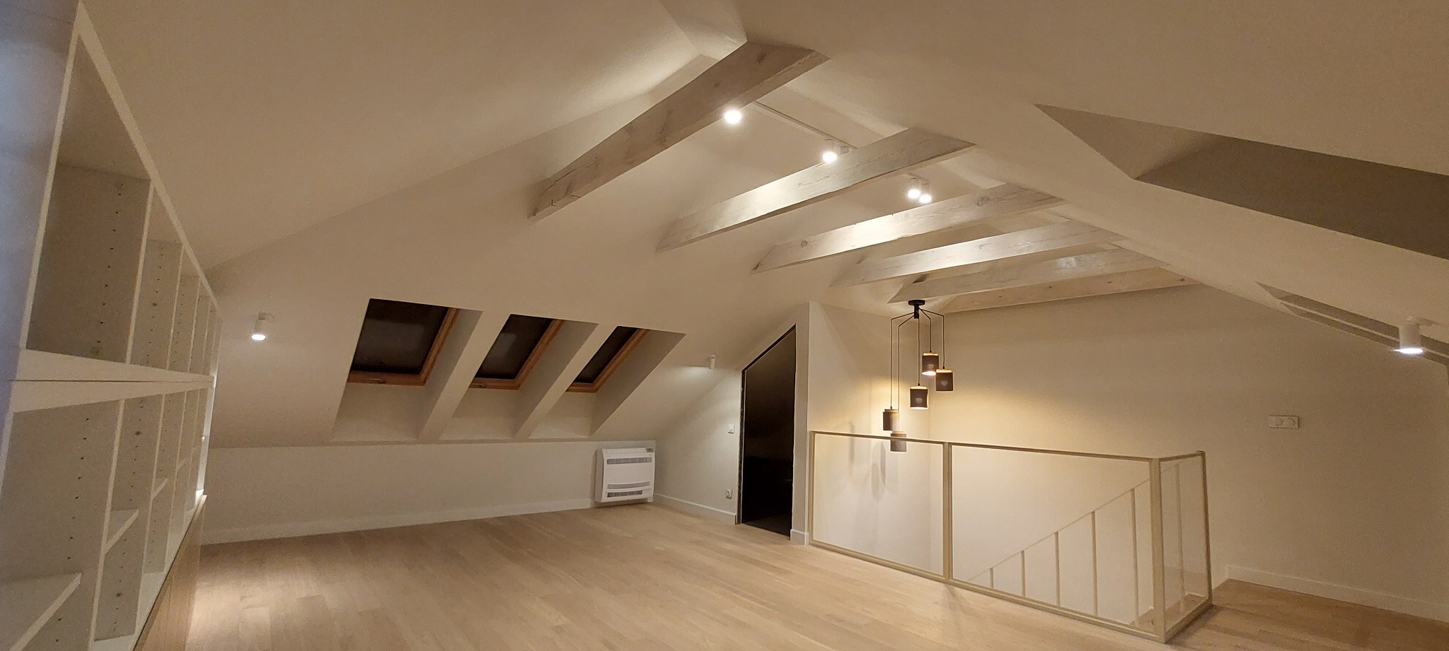 Jasne poddasze z drewnianą podłogą, skośnym sufitem z belkami i oknami dachowymi, oraz nowoczesnym oświetleniem i regałem.