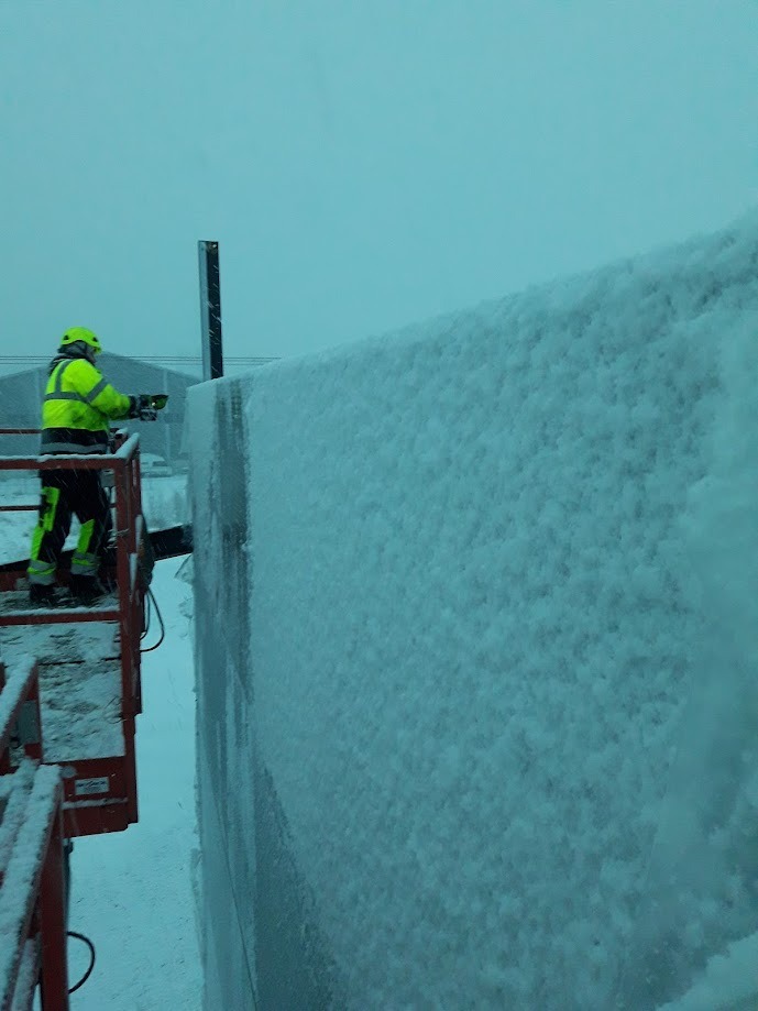 Pracownik w żółtym kombinezonie na podnośniku montuje płytę warstwową na hali w zimowych warunkach, widoczny fragment konstrukcji stalowej.