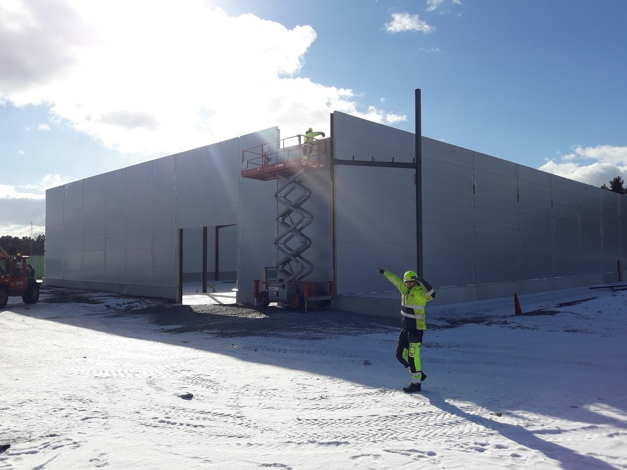 Pracownik w odblaskowym ubraniu wskazuje na budowę hali z płyt warstwowych, drugi pracownik na podnośniku koszowym montuje elementy konstrukcji, zimowy krajobraz.