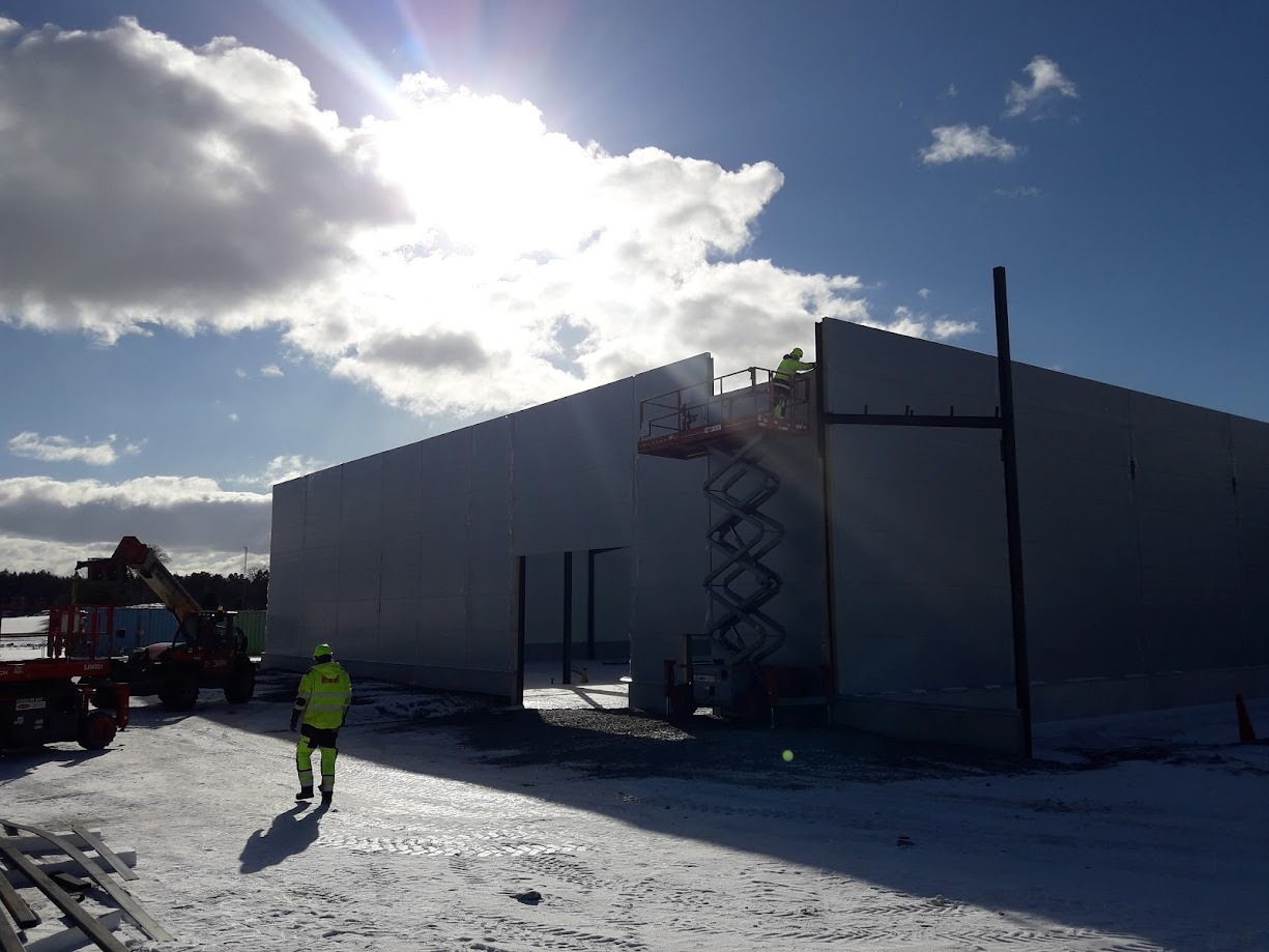 Pracownicy w odblaskowych ubraniach montują pionowe płyty na konstrukcji hali, jeden na podnośniku nożycowym, drugi idzie po śniegu, w tle widoczne maszyny budowlane i pochmurne niebo.