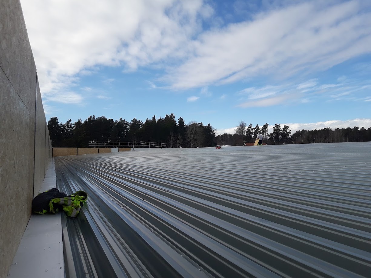 Widok z góry na rozległy dach z blachy trapezowej w trakcie budowy, z widoczną ścianą boczną z płyty OSB i pozostawioną odzieżą roboczą w rogu, na tle błękitnego nieba z chmurami i odległego lasu.