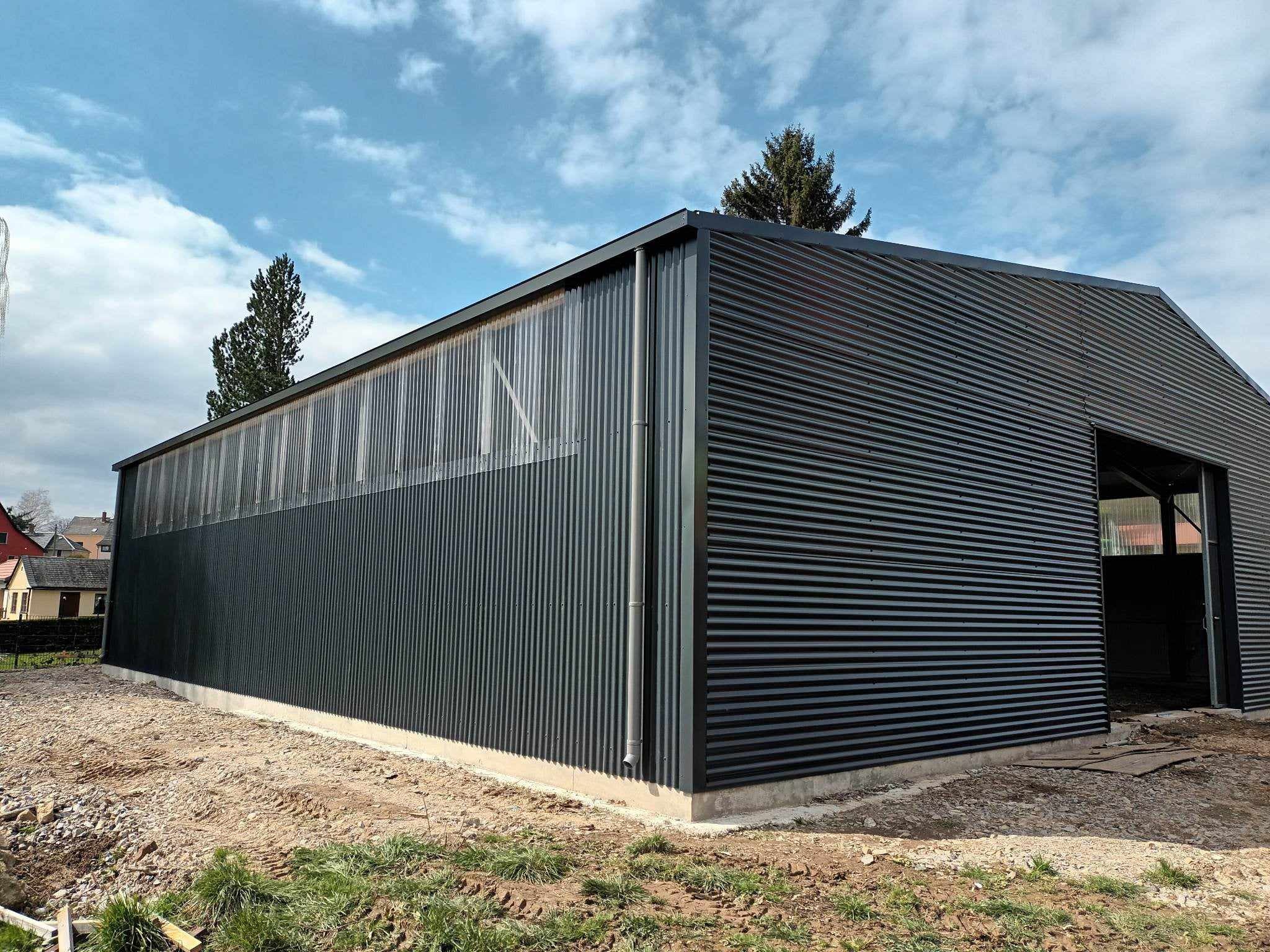 Ciemna hala przemysłowa z blachy falistej, częściowo z panelami transparentnymi, na placu budowy z widocznymi śladami opon i resztkami materiałów.